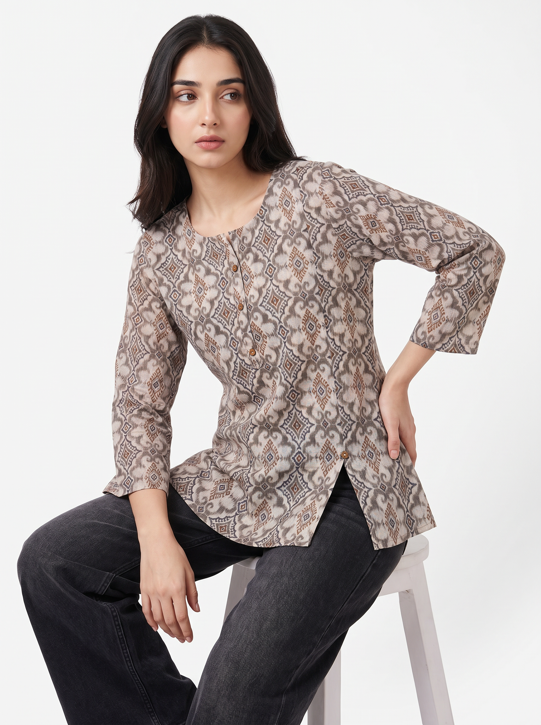 A woman seated on a white stool wearing a long-sleeve button-front tunic top with an allover abstract floral pattern in beige, taupe, and brown tones, paired with dark gray jeans against a plain light background.