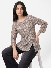 A woman seated on a white stool wearing a long-sleeve button-front tunic top with an allover abstract floral pattern in beige, taupe, and brown tones, paired with dark gray jeans against a plain light background.