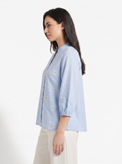 A woman shown in side profile wearing a light blue button-front blouse with small yellow and white floral embroidery and cream pants, photographed against a plain white studio background.