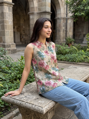 A woman sitting on a stone bench in a garden courtyard, wearing a sleeveless beige top with a pink and green floral print and light blue jeans.