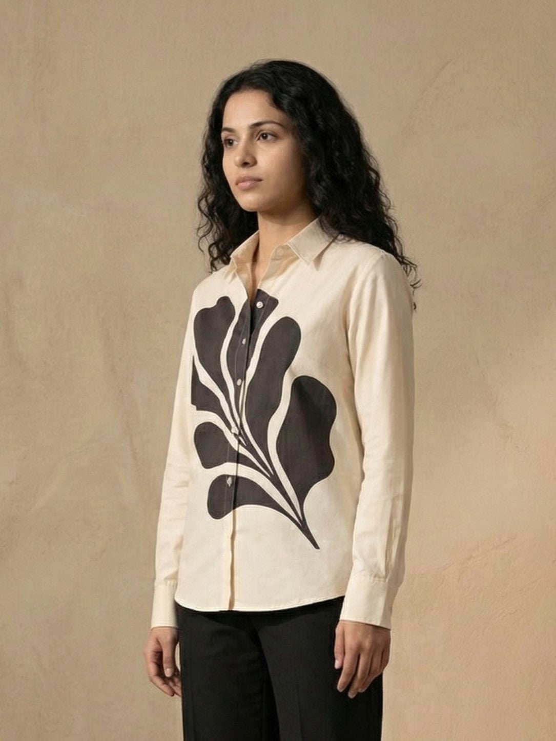 A woman modeling a cream-colored long-sleeve button-up shirt with a large black abstract leaf pattern on the front, paired with dark gray trousers, set against a textured beige background.