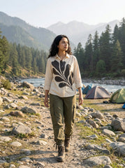 A woman modeling a cream-colored long-sleeve button-up shirt with a large black abstract leaf pattern on the front, paired with dark gray trousers, set against a textured beige background.