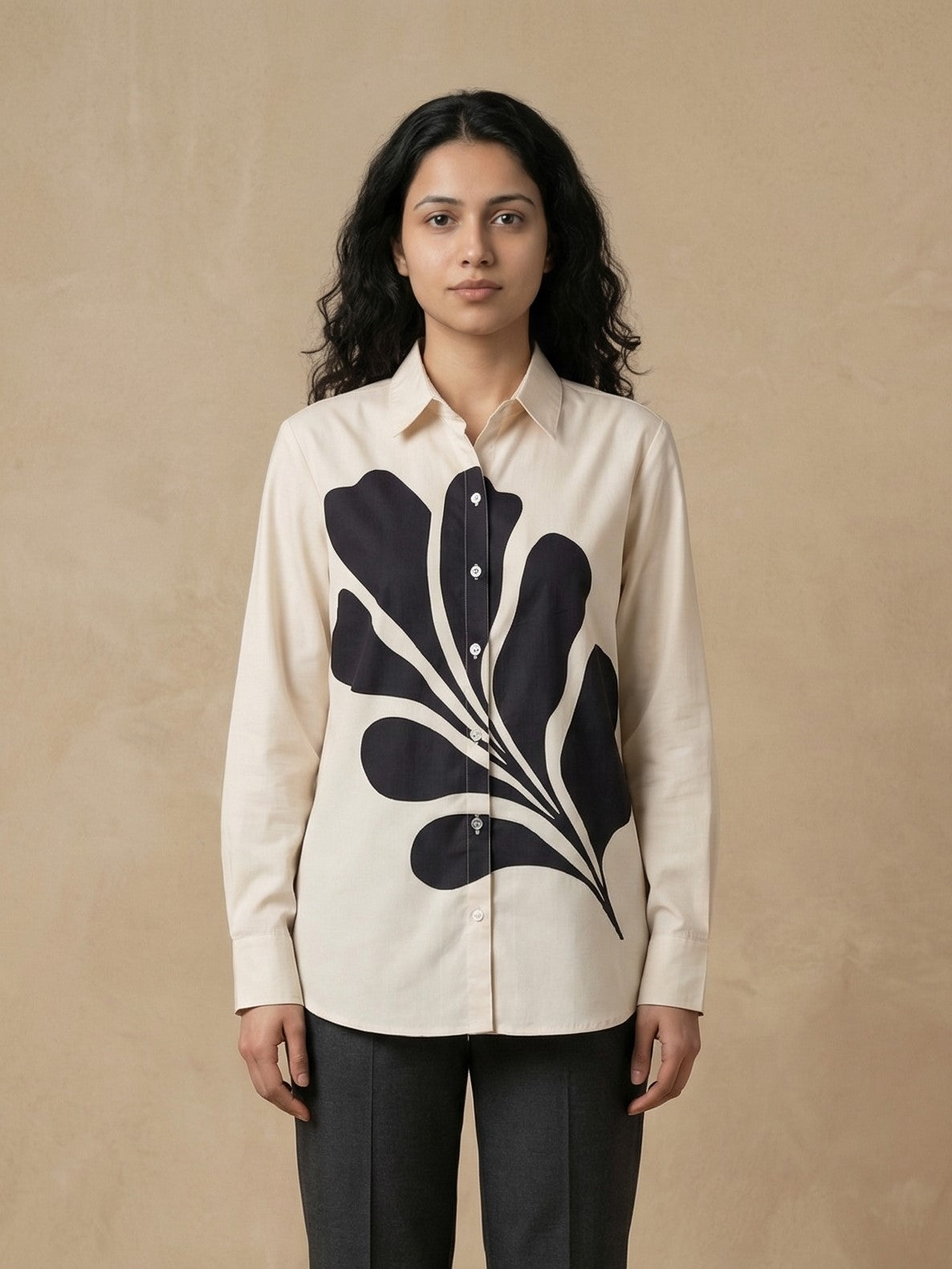 A woman modeling a cream-colored long-sleeve button-up shirt with a large black abstract leaf pattern on the front, paired with dark gray trousers, set against a textured beige background.
