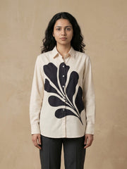 A woman modeling a cream-colored long-sleeve button-up shirt with a large black abstract leaf pattern on the front, paired with dark gray trousers, set against a textured beige background.