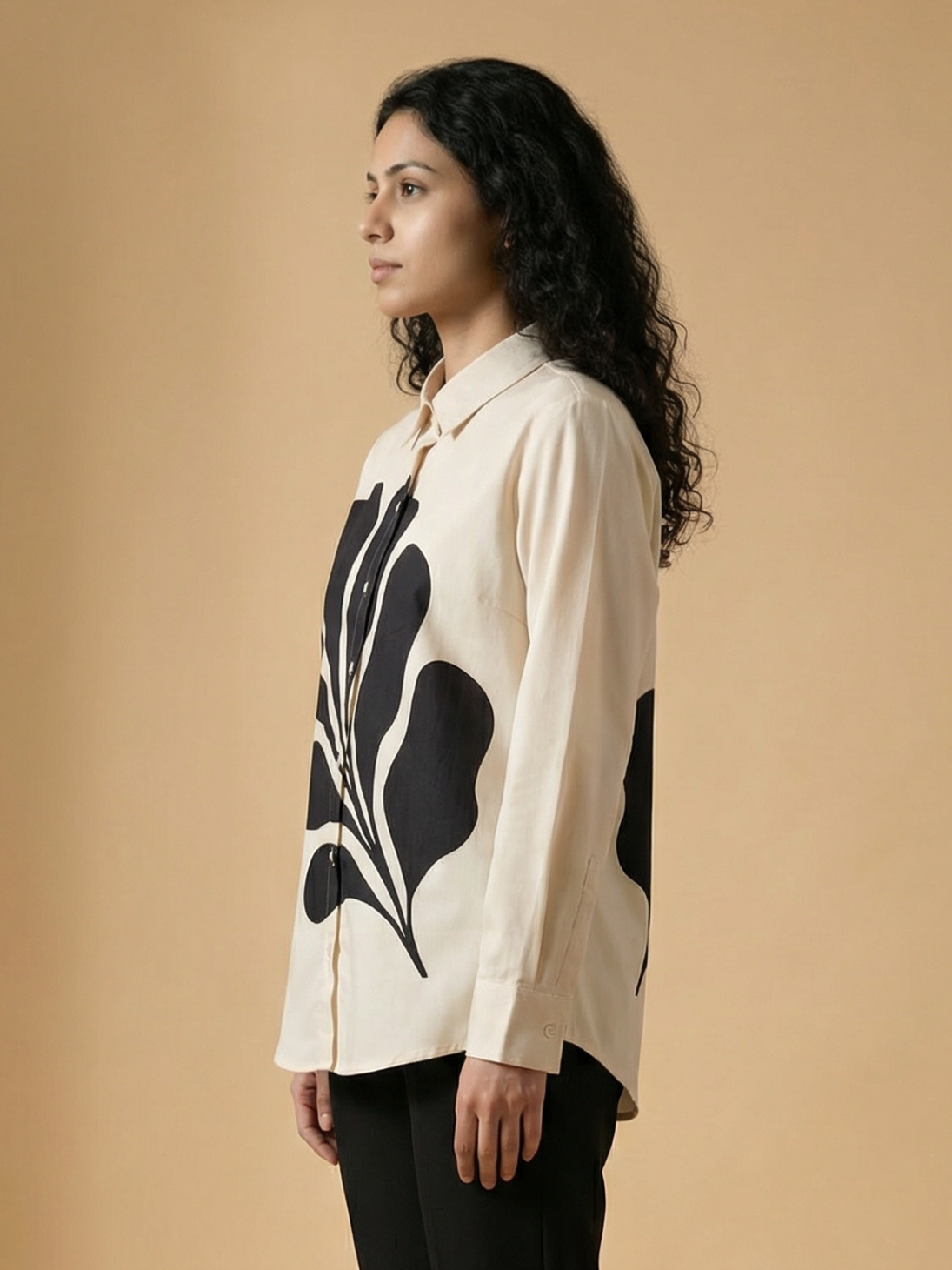 A woman with long curly black hair is shown in profile wearing a cream-colored long-sleeve button-up shirt with a large black abstract leaf-like print, paired with black pants, against a solid beige studio background.