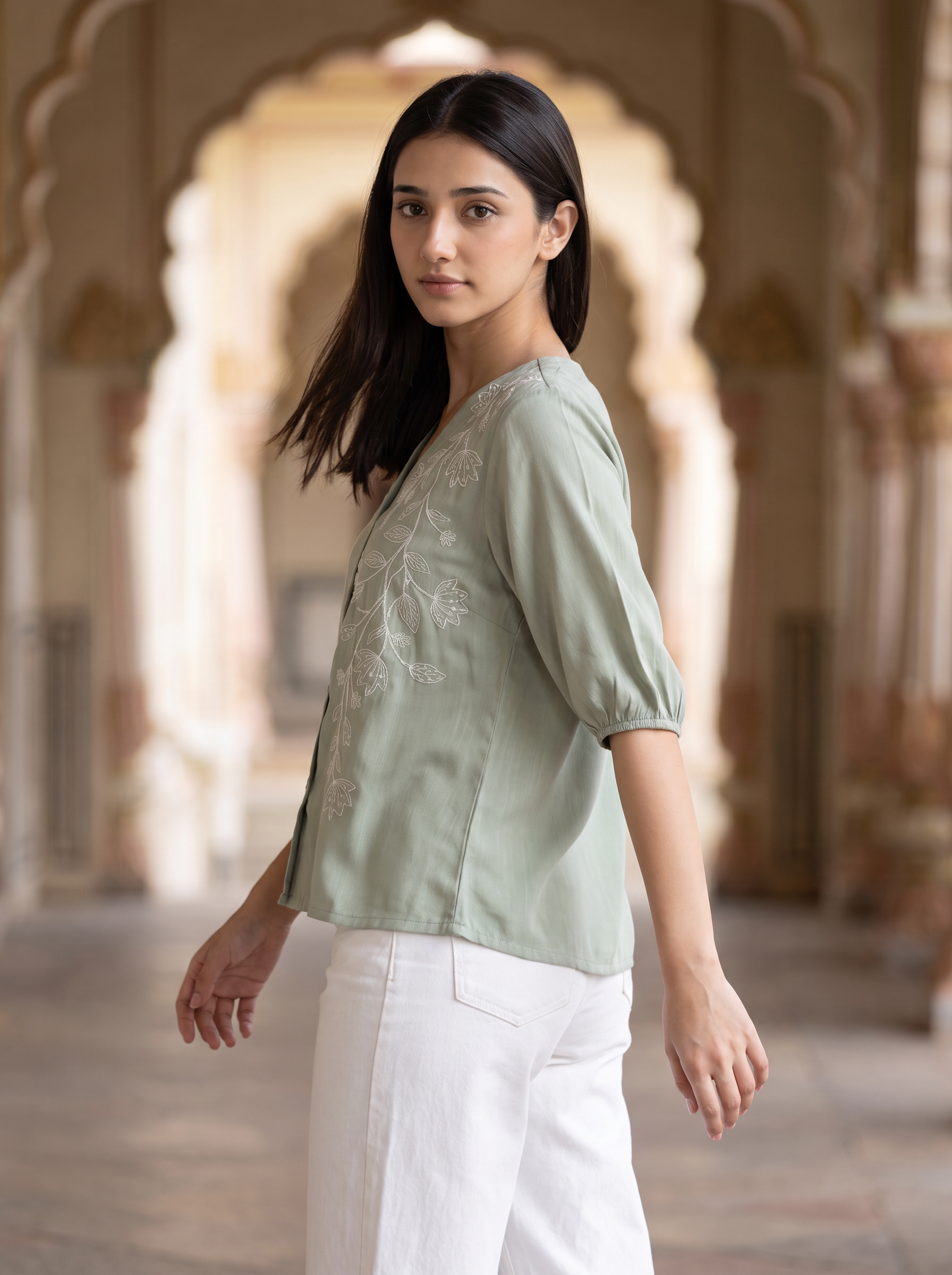 A woman wearing a sage green embroidered blouse with white pants, standing in a historic stone corridor with arches.