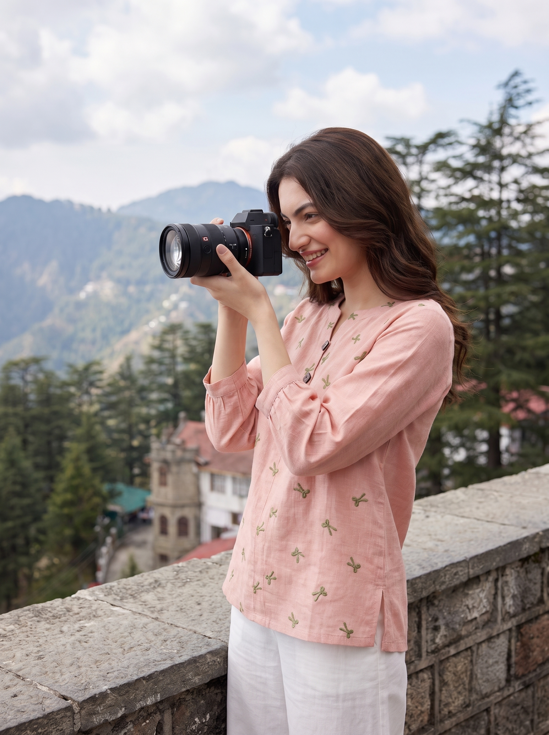 A woman holding a camera stands outdoors on a stone ledge with mountains and trees in the background, wearing a light pink embroidered top and white pants.