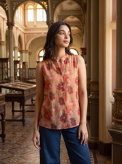 A woman wearing a sleeveless button-up blouse with a floral print in peach, rust, and olive tones, paired with dark blue pants, standing in an ornate indoor architectural setting with arches and columns.