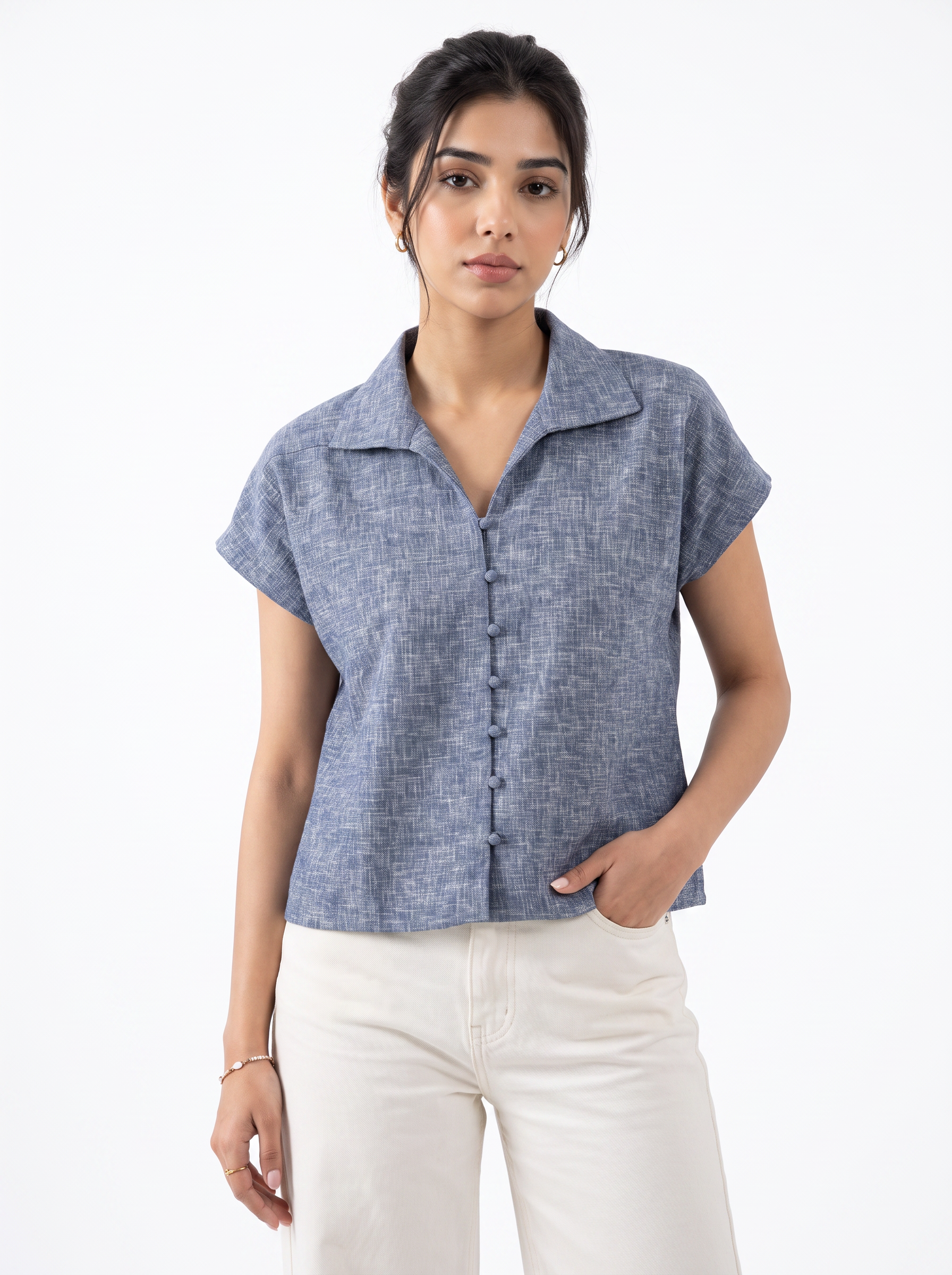 A woman modeling a short-sleeve button-front collared top in a heathered blue-gray fabric, paired with off-white pants, against a plain white studio background.