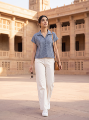 A woman walking outdoors in front of a sandstone architectural courtyard, wearing a short-sleeve button-front collared top in a muted blue-gray, off-white wide-leg pants, white sneakers, and carrying a brown shoulder bag.