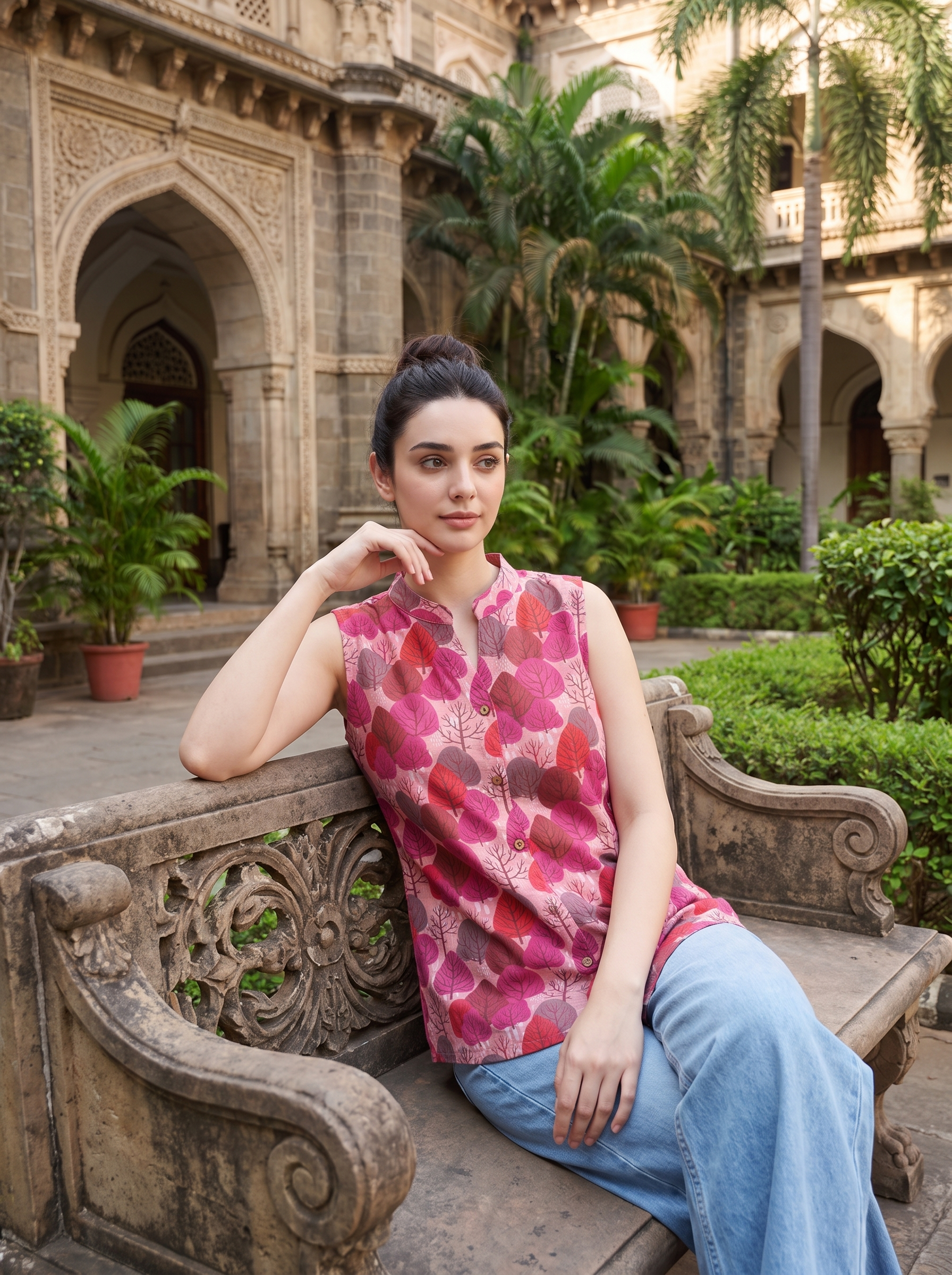 A woman sits on an ornate stone bench in a courtyard, wearing a sleeveless pink and red floral print top with light blue jeans. The background features historic stone architecture, palm trees, and green plants.