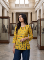 A woman stands in a grand museum or gallery wearing a yellow three-quarter sleeve kurta with a black and red geometric pattern, paired with dark blue jeans.