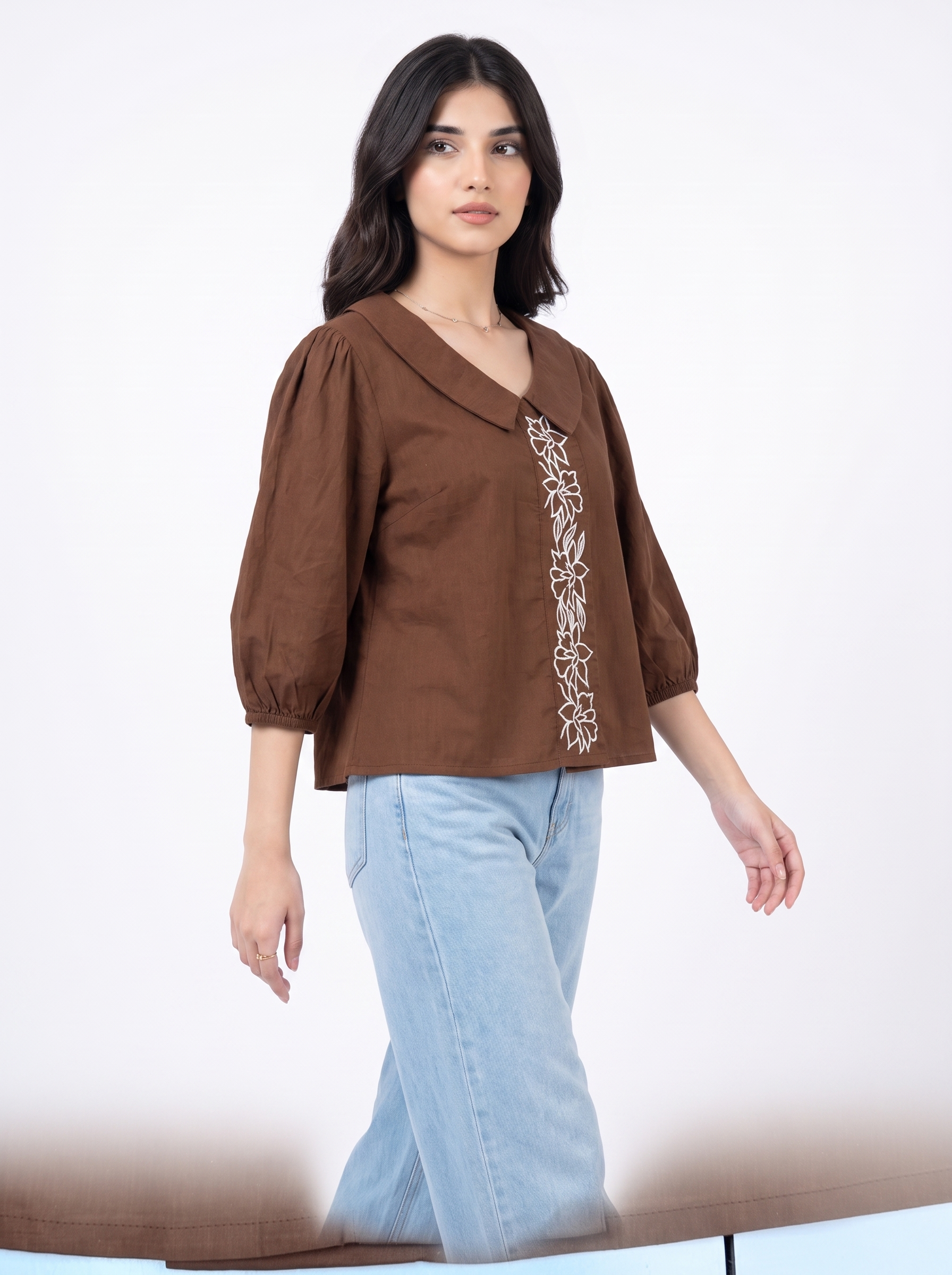 A woman modeling a brown three-quarter sleeve blouse with vertical white floral embroidery on the front, paired with light blue jeans, against a plain white studio background.