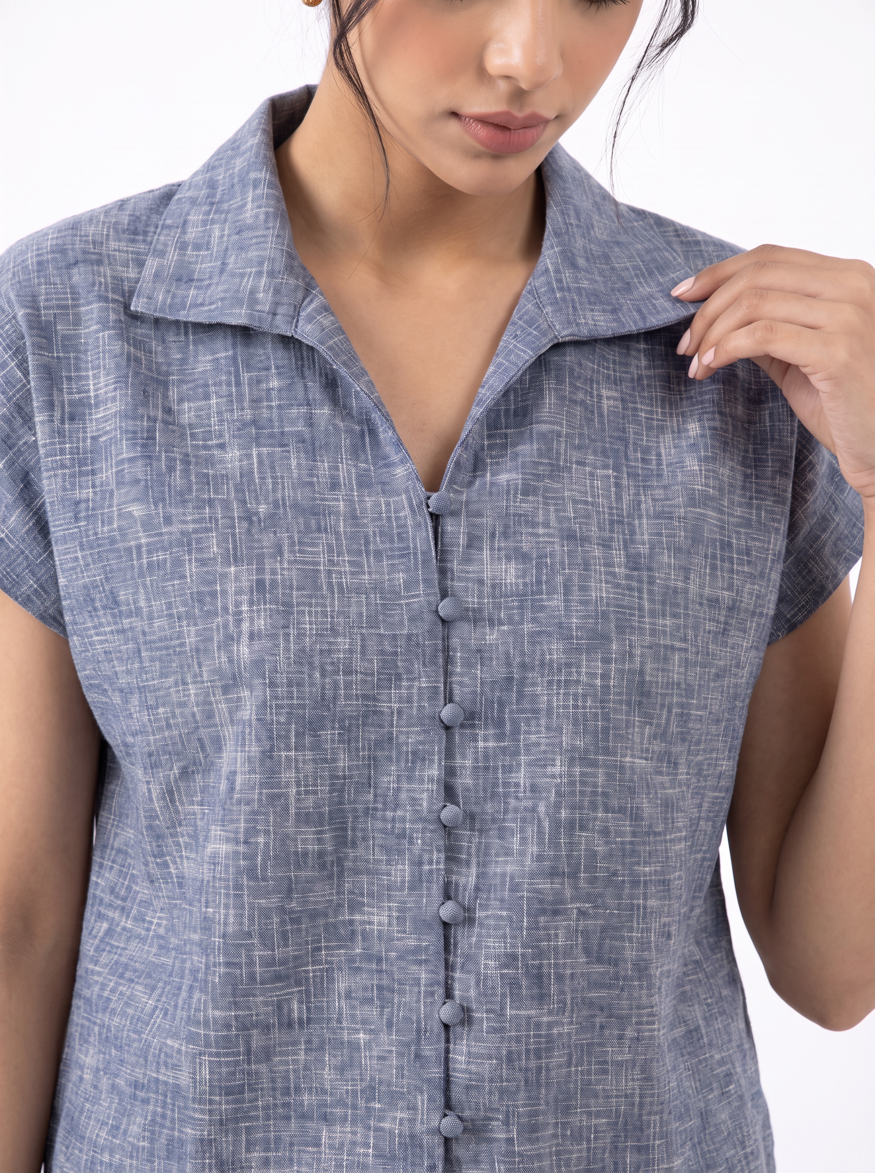 A close-up of a woman wearing a short-sleeve button-front top with a textured, heathered blue-gray fabric and a pointed collar.