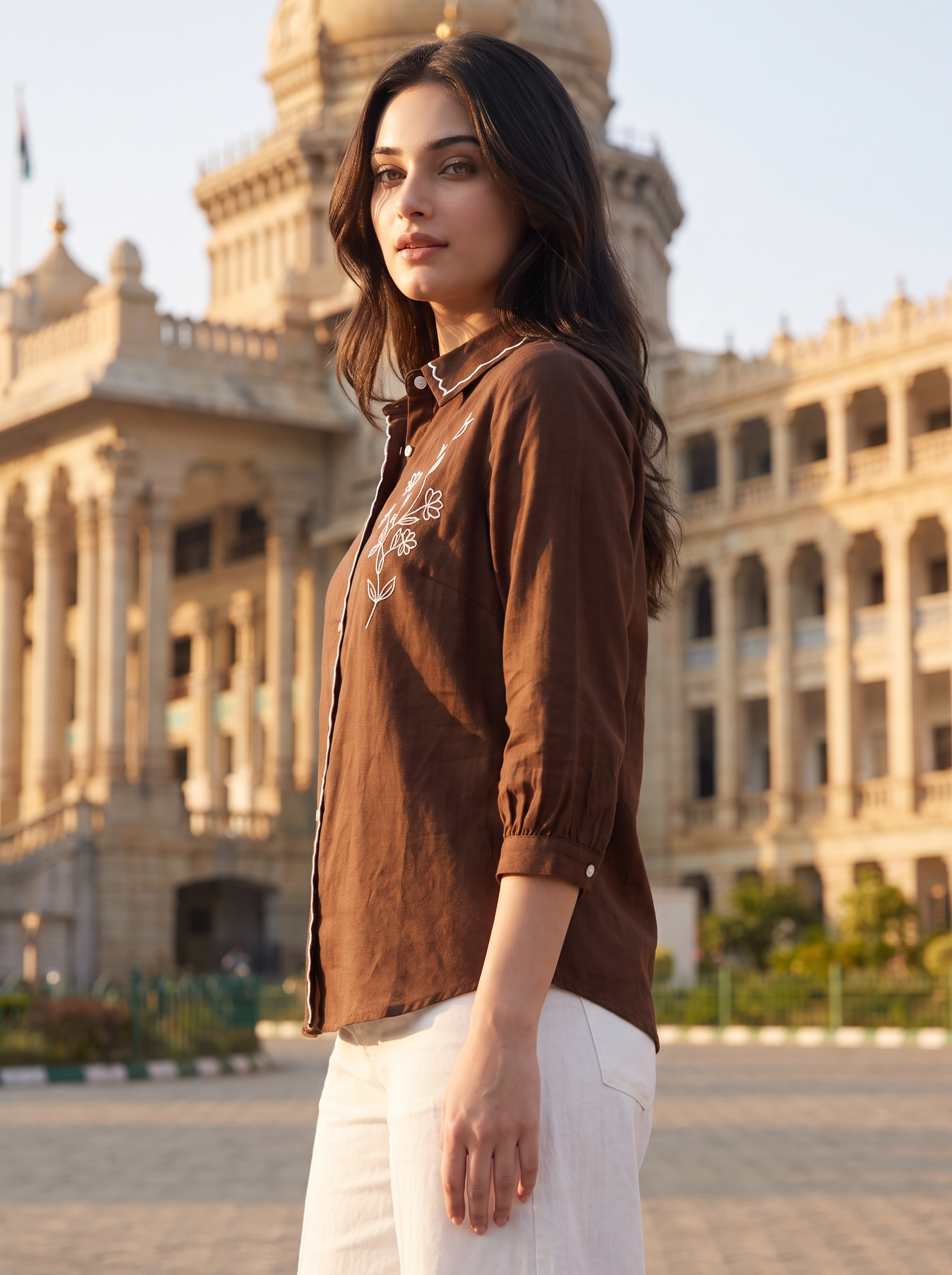 A woman wearing a brown long-sleeve button-up shirt with white pants, standing in front of a large historic building.