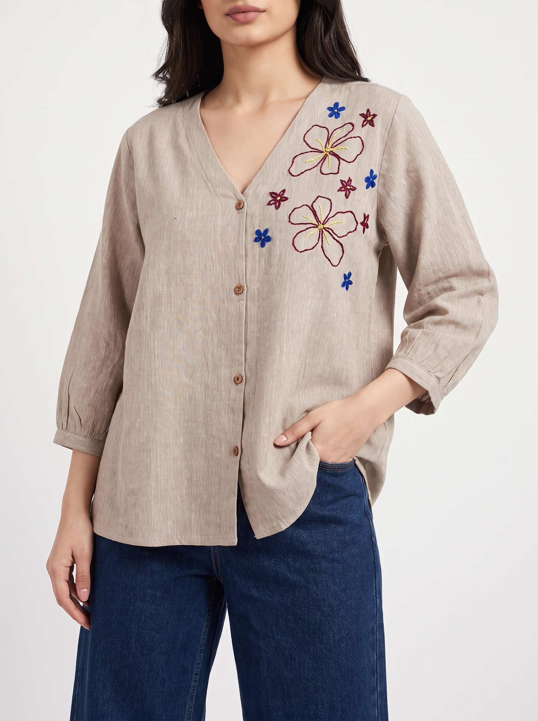 A woman wearing a beige linen button-up blouse with colorful floral embroidery on the chest and dark blue jeans, photographed against a clean white studio background.