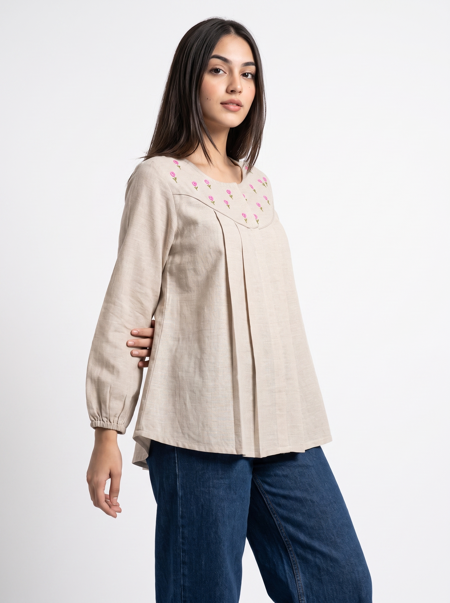 A woman modeling a light beige linen long-sleeve top with pink floral embroidery around the round neckline, paired with blue jeans, against a plain white studio background.