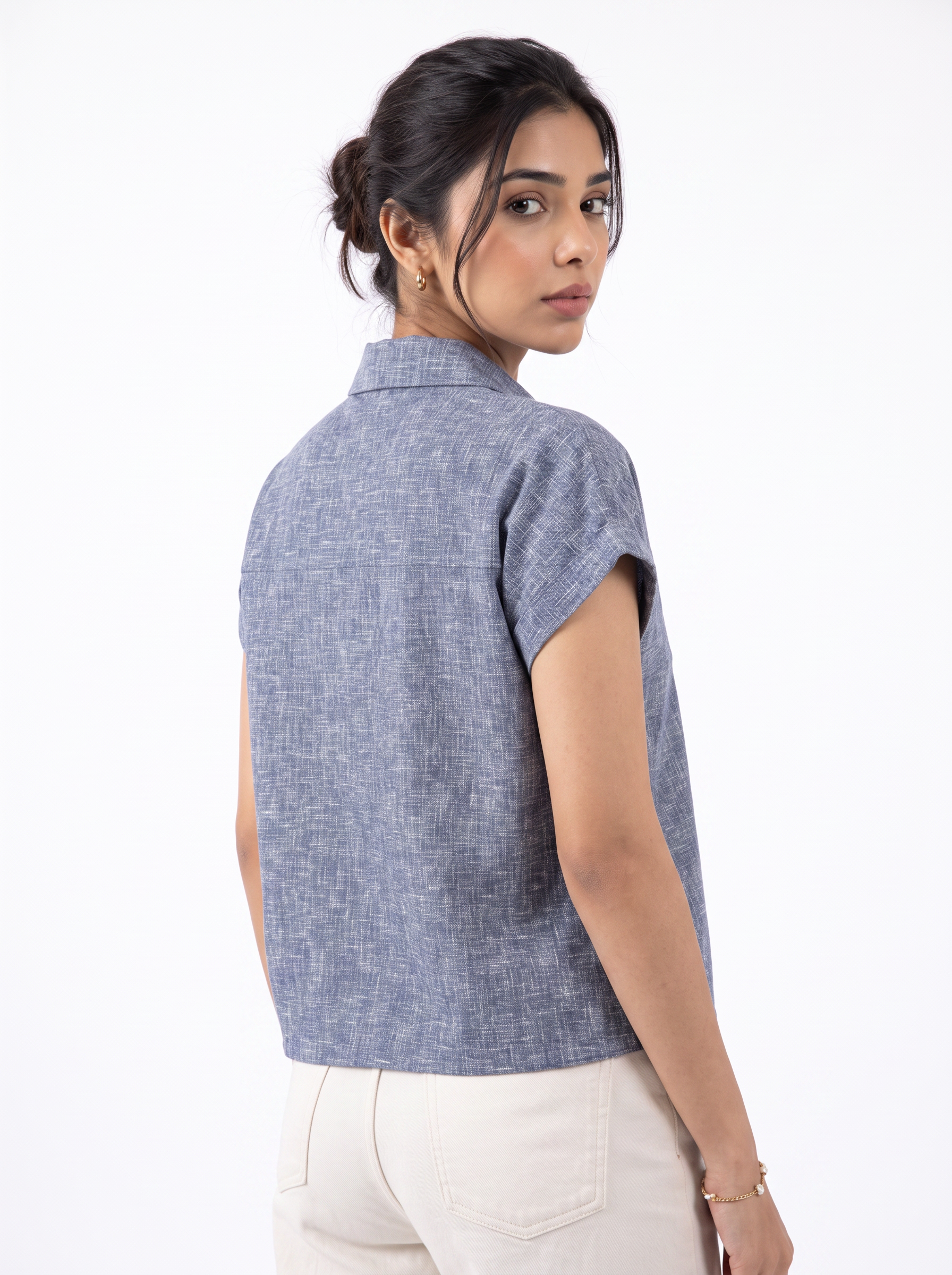 A woman shown from the back wearing a textured slate blue short-sleeve collared top and cream-colored pants, photographed against a clean white studio background.