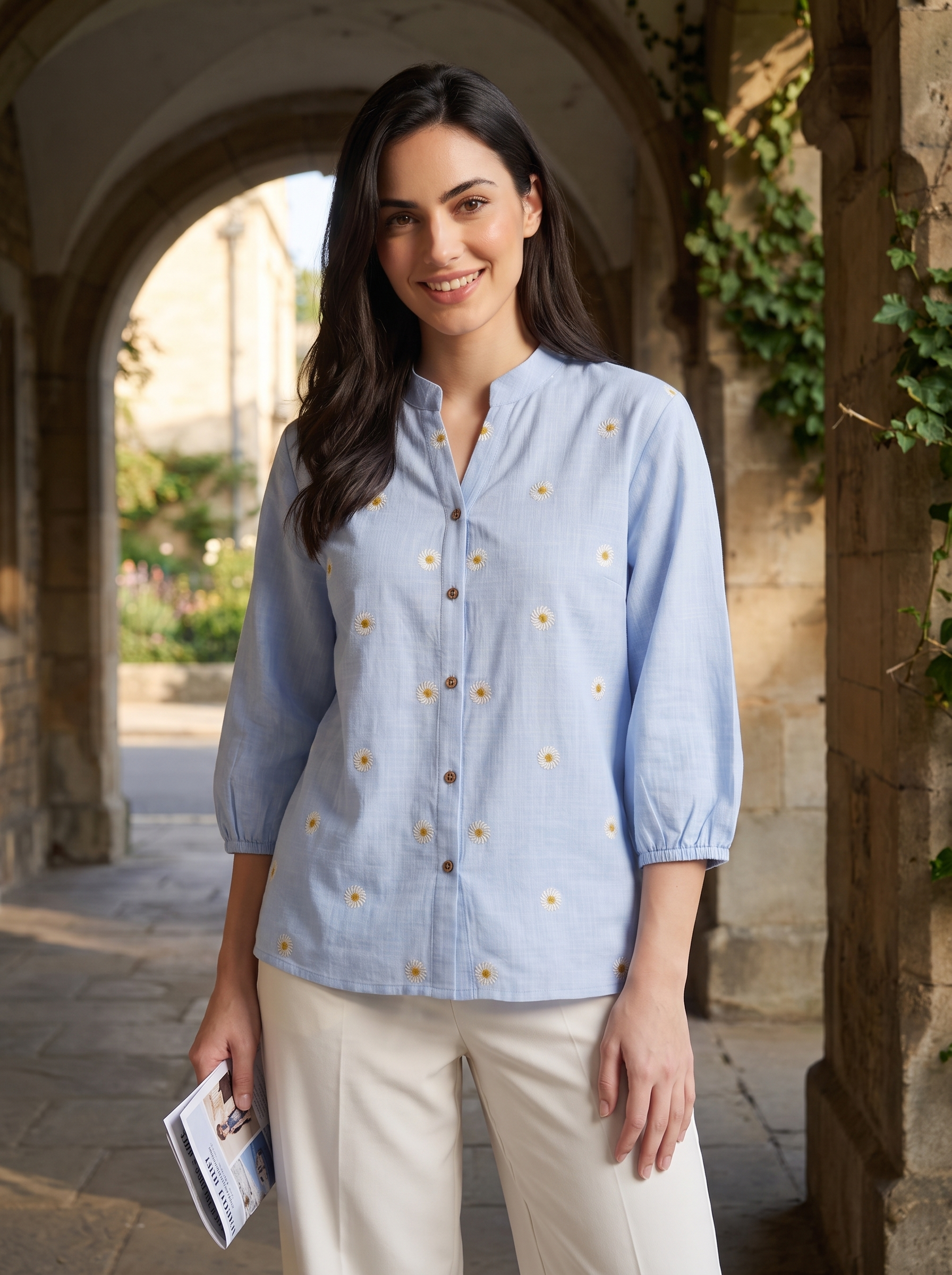A woman stands in a stone archway corridor wearing a light blue button-front blouse with small embroidered floral details and cream pants, holding a brochure.
