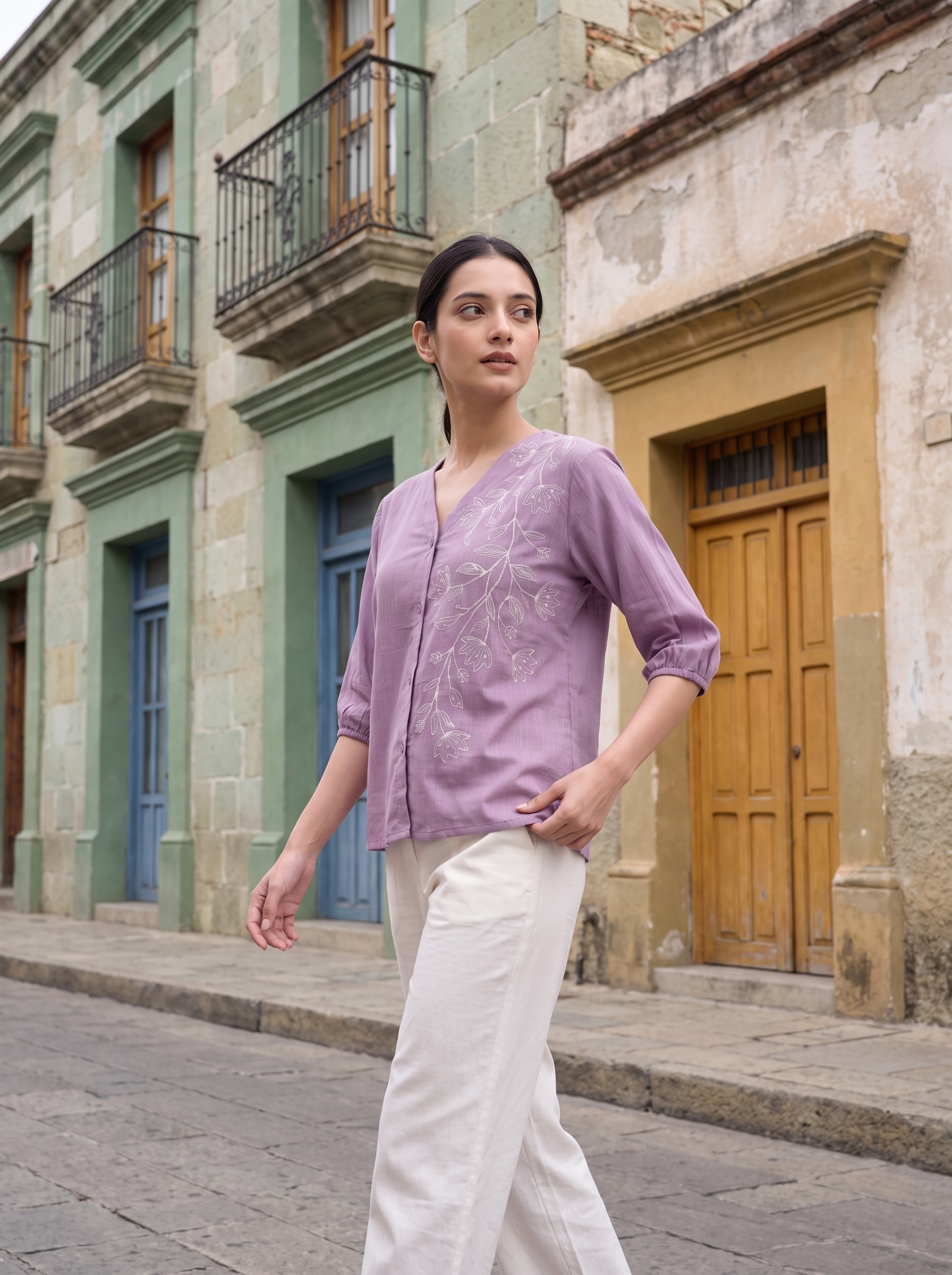 A woman stands on a cobblestone street wearing a lavender embroidered blouse and white trousers, with historic stone buildings featuring green and blue doors in the background.
