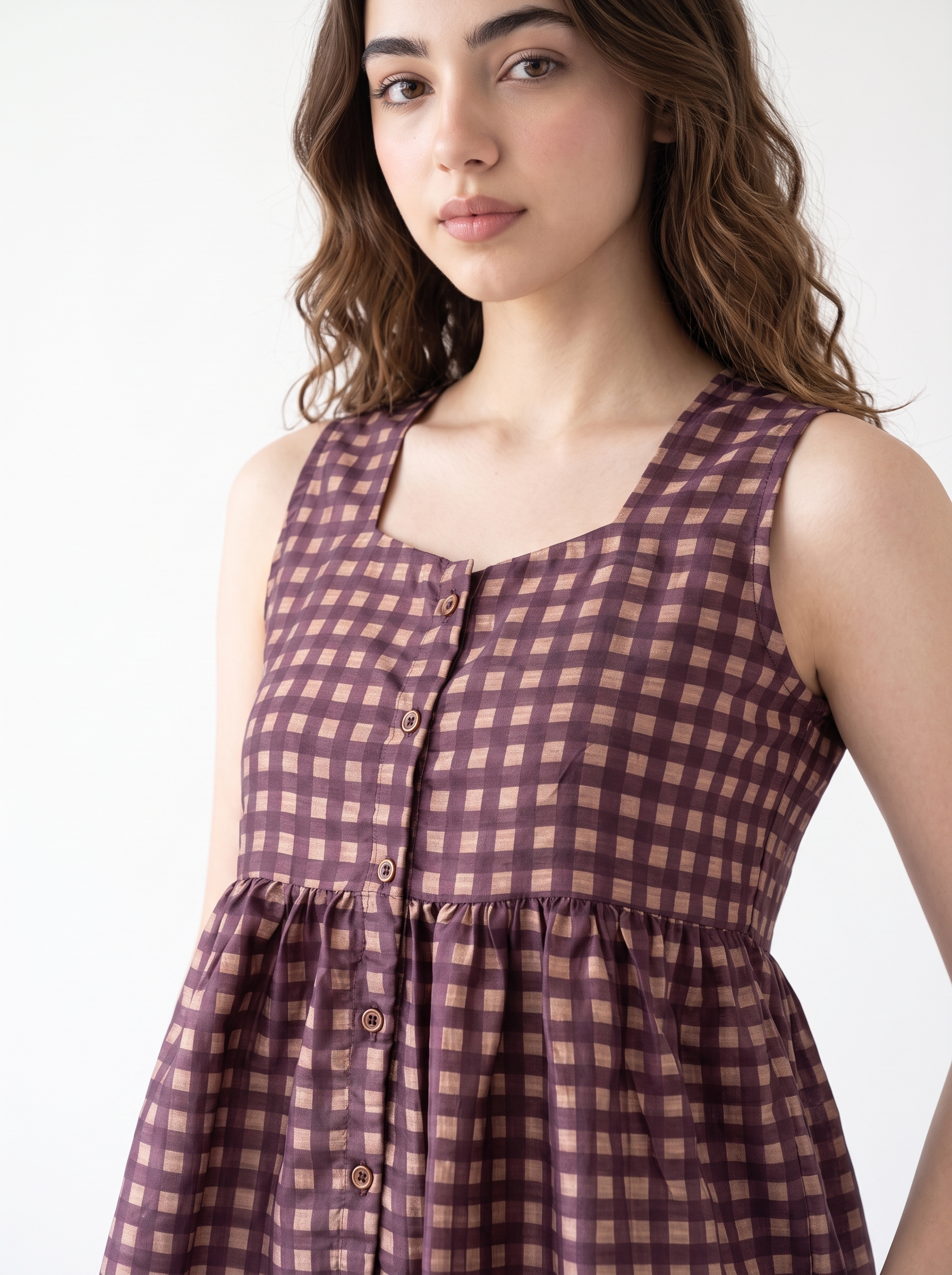 A woman wearing a sleeveless button-front dress with a brown and dusty rose gingham pattern, shown against a plain white background.