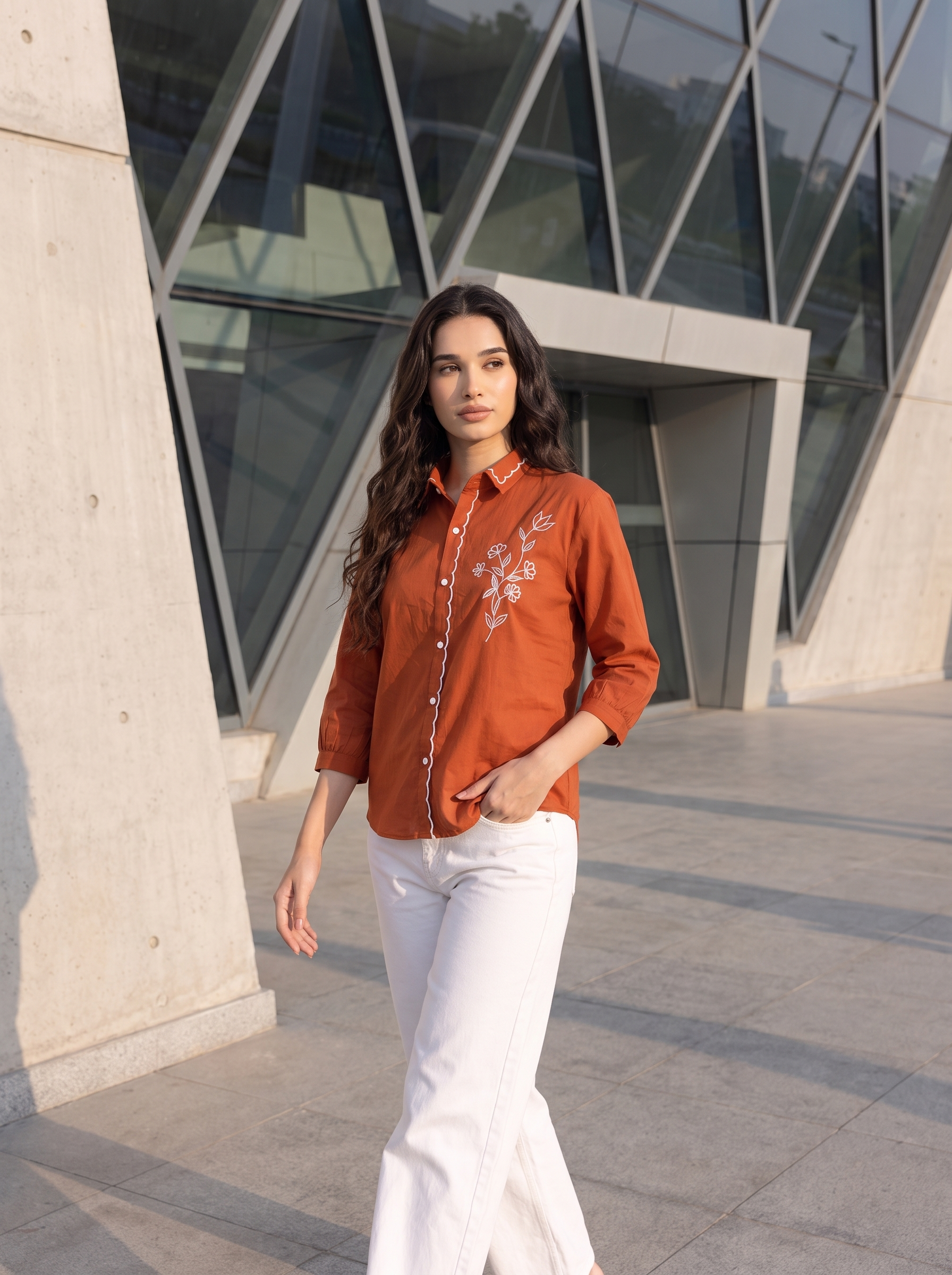 A woman wearing a rust-orange button-up shirt with white floral embroidery on the chest and white pants, standing outdoors in front of a modern glass and concrete building.