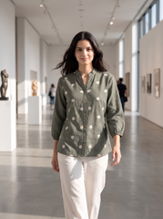 A woman walking down a bright, modern art gallery wearing a sage green linen button-up shirt with white floral prints and white pants.