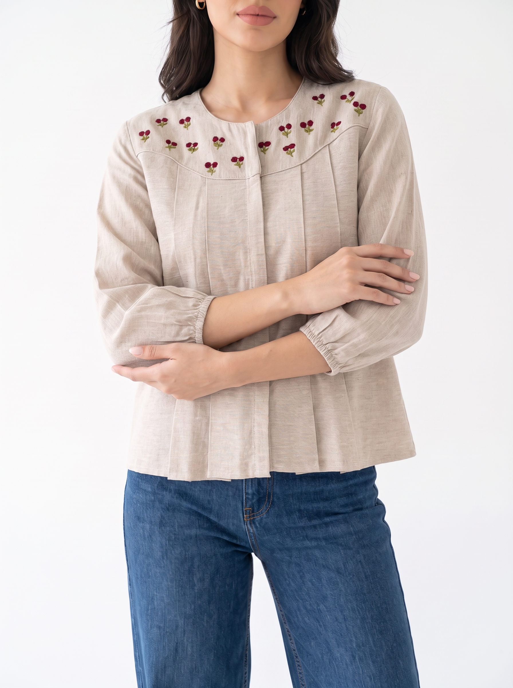 A woman wearing a beige linen blouse with embroidered cherry details on the yoke and blue jeans, standing against a plain white background.