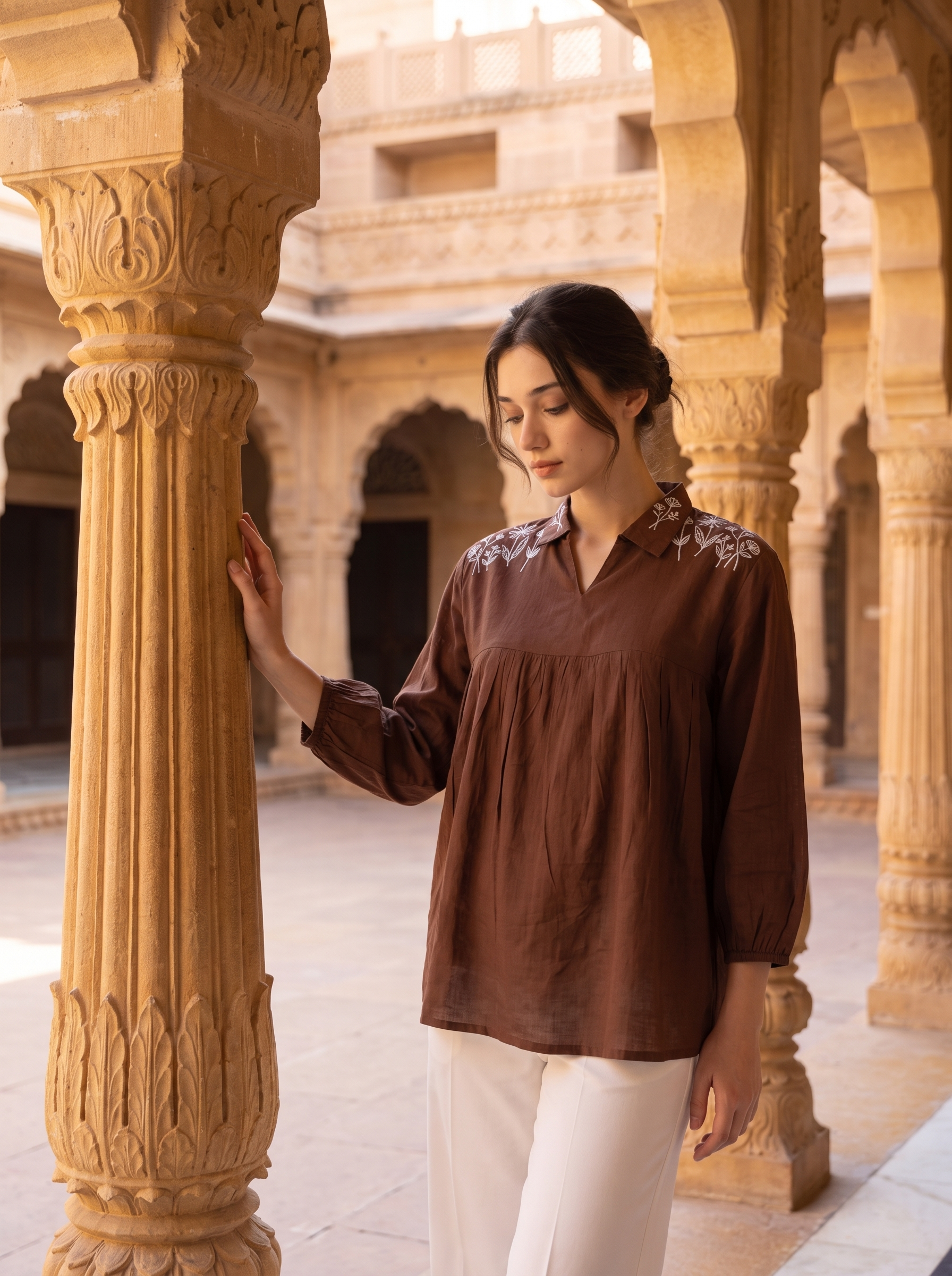 A woman wearing a brown long-sleeve blouse with embroidered details on the shoulders and white pants, standing in a sunlit stone courtyard with ornate columns.