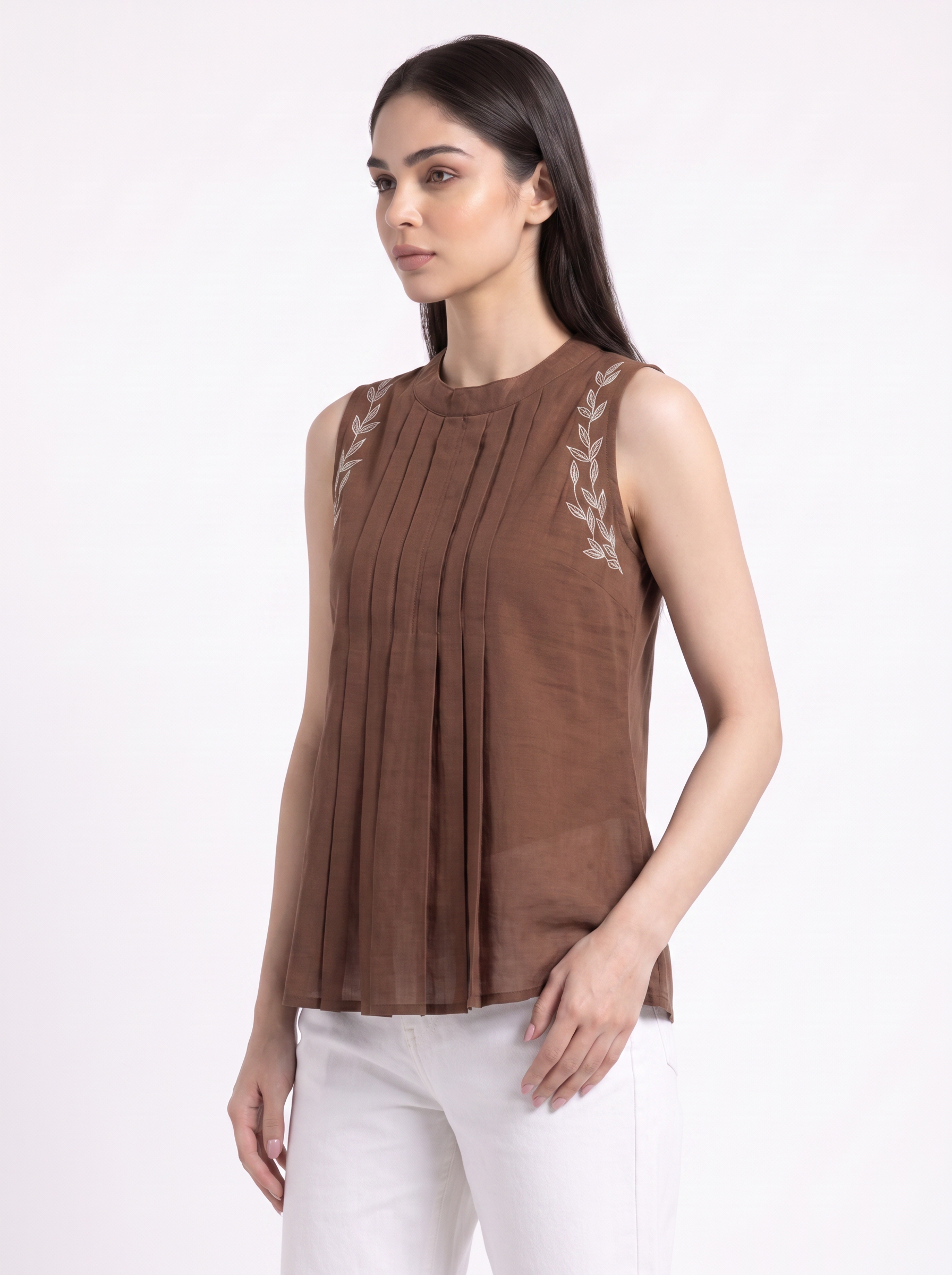 A woman modeling a sleeveless brown pleated top with white embroidered leaf details on the shoulders, paired with white pants, against a plain white background.