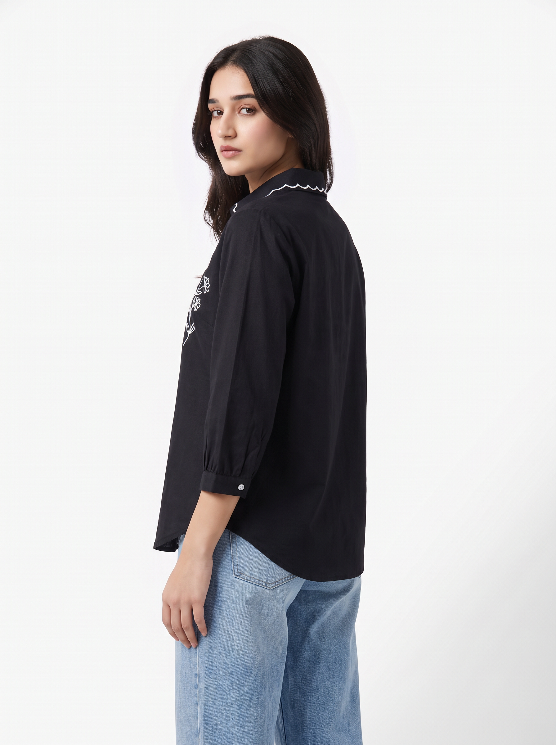 A woman modeling a black long-sleeve button-up shirt with white embroidered details on the chest and collar, paired with light blue jeans, against a plain white studio background.