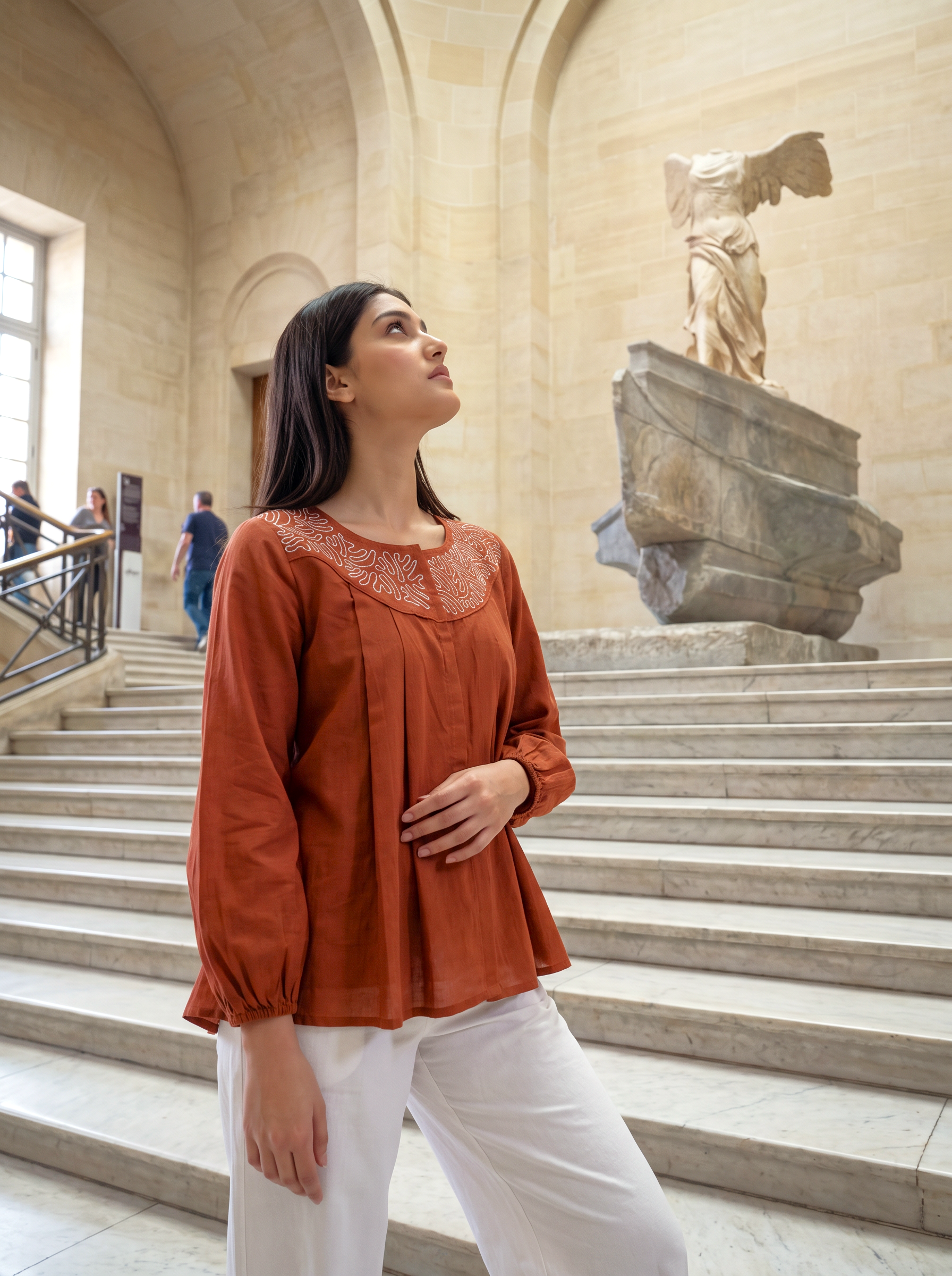 A woman wearing a rust-orange long-sleeve blouse with a lace V-neckline and white pants stands on marble stairs inside a grand classical museum hall, looking upward toward a large stone statue.