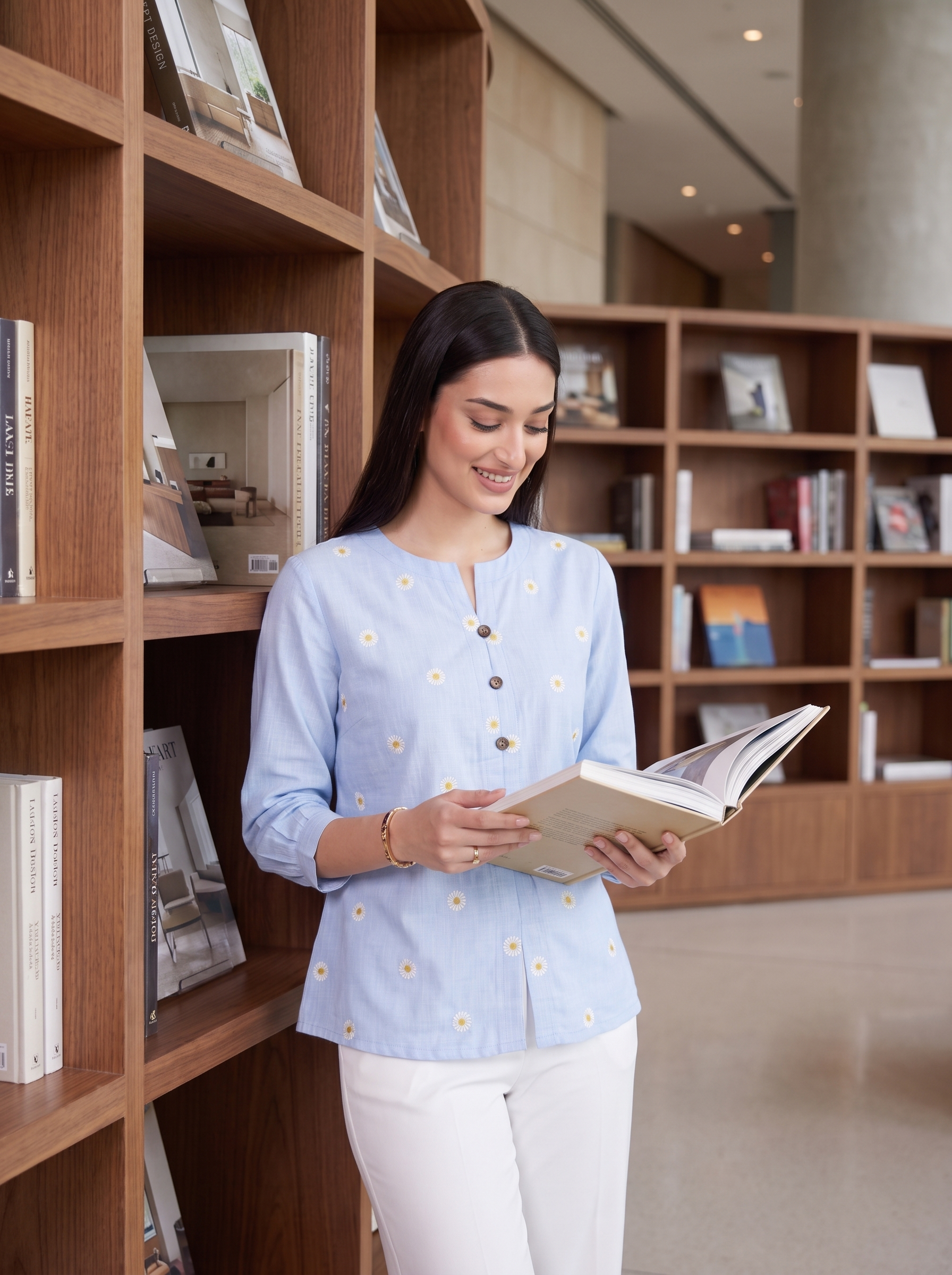 A woman in a light blue floral printed kurta and white trousers stands in a library, reading an open book.