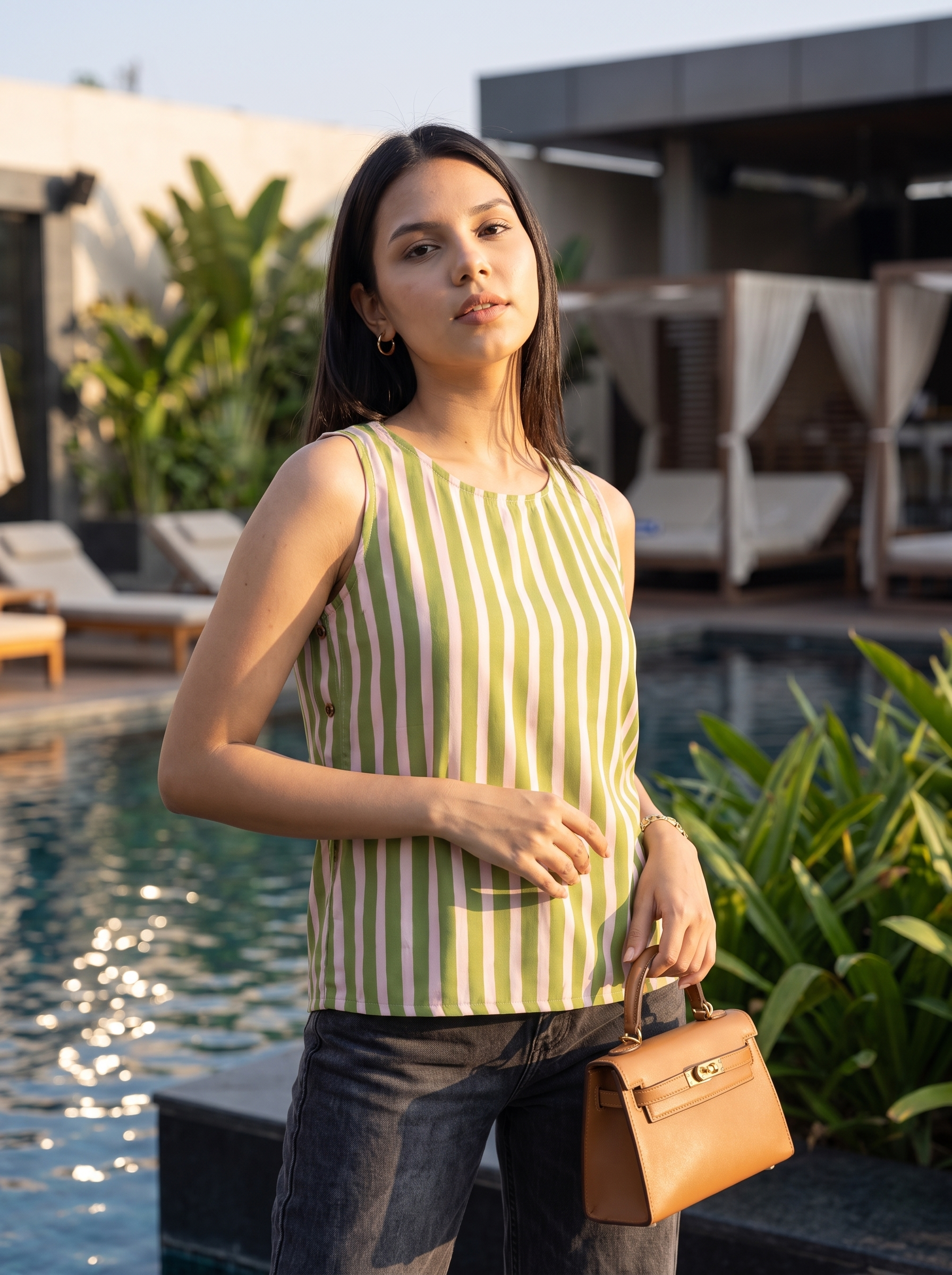 A woman stands by a swimming pool wearing a sleeveless top with vertical lime green and white stripes, paired with dark jeans and holding a tan handbag.