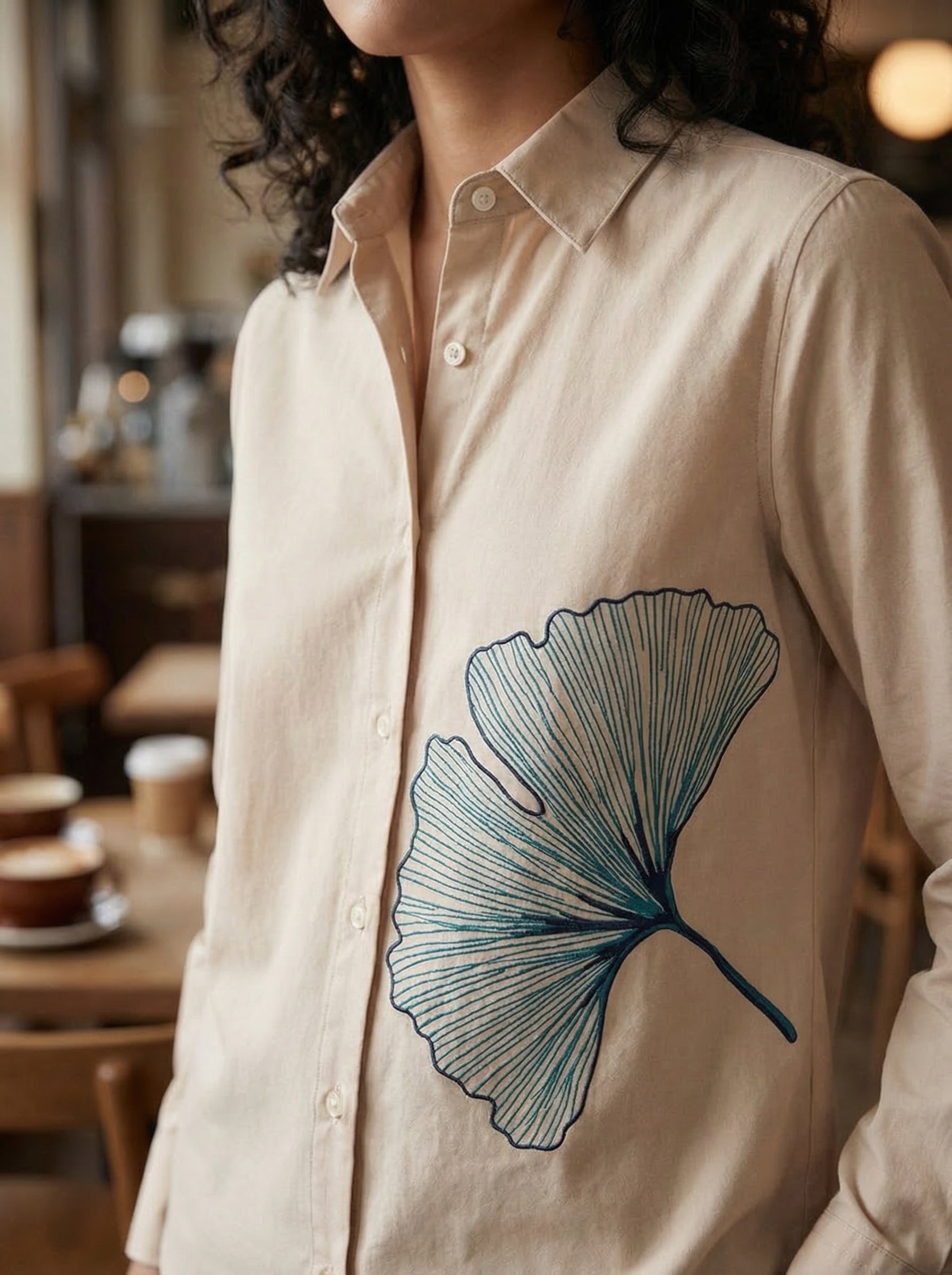 A refined cream shirt top featuring a striking teal ginkgo leaf graphic on the front for a clean artistic look.