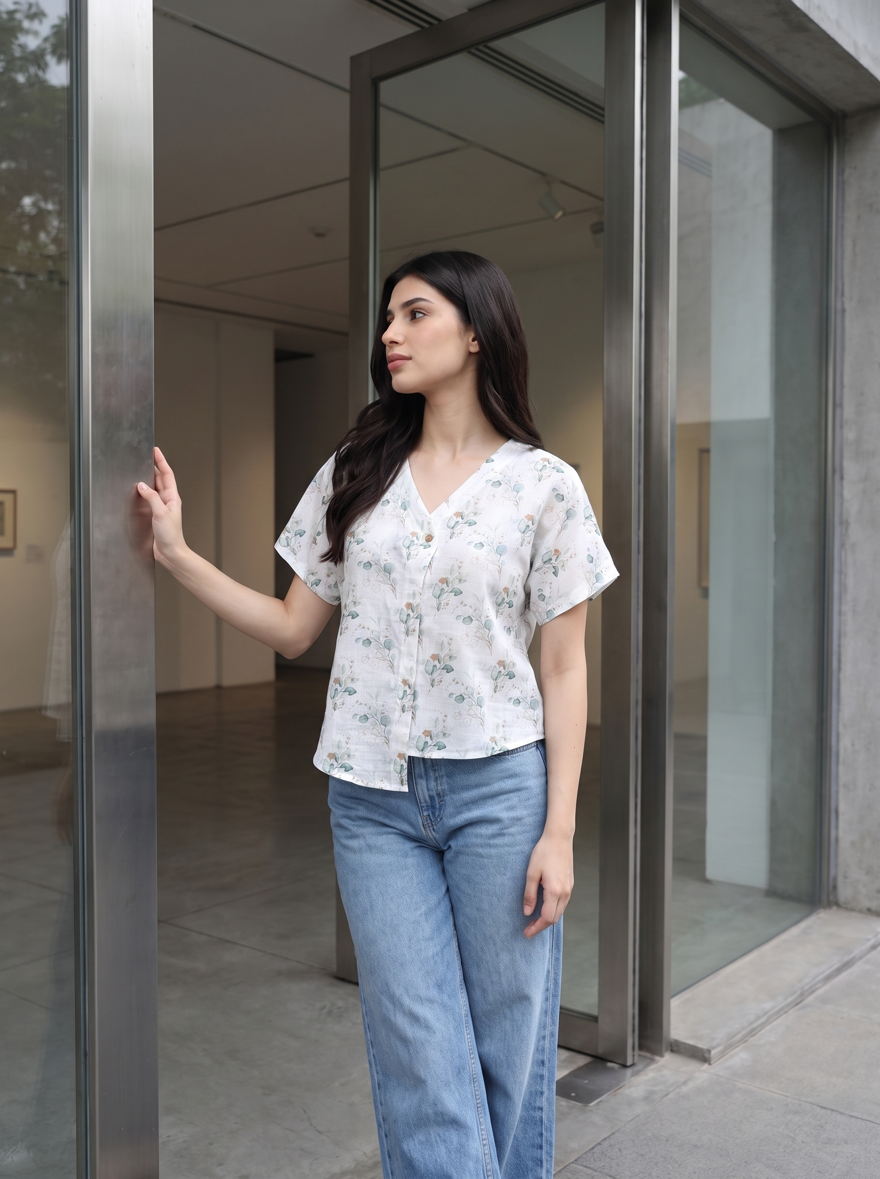 A woman wearing a white short-sleeve button-up blouse with a delicate blue and green floral print, paired with light-wash blue jeans, standing in a modern glass entryway.