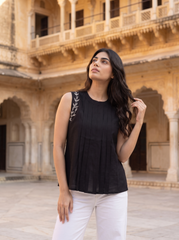 A woman wearing a black sleeveless pleated top with silver embroidery on the shoulder and white pants, standing in a courtyard with a historic sandstone building in the background.