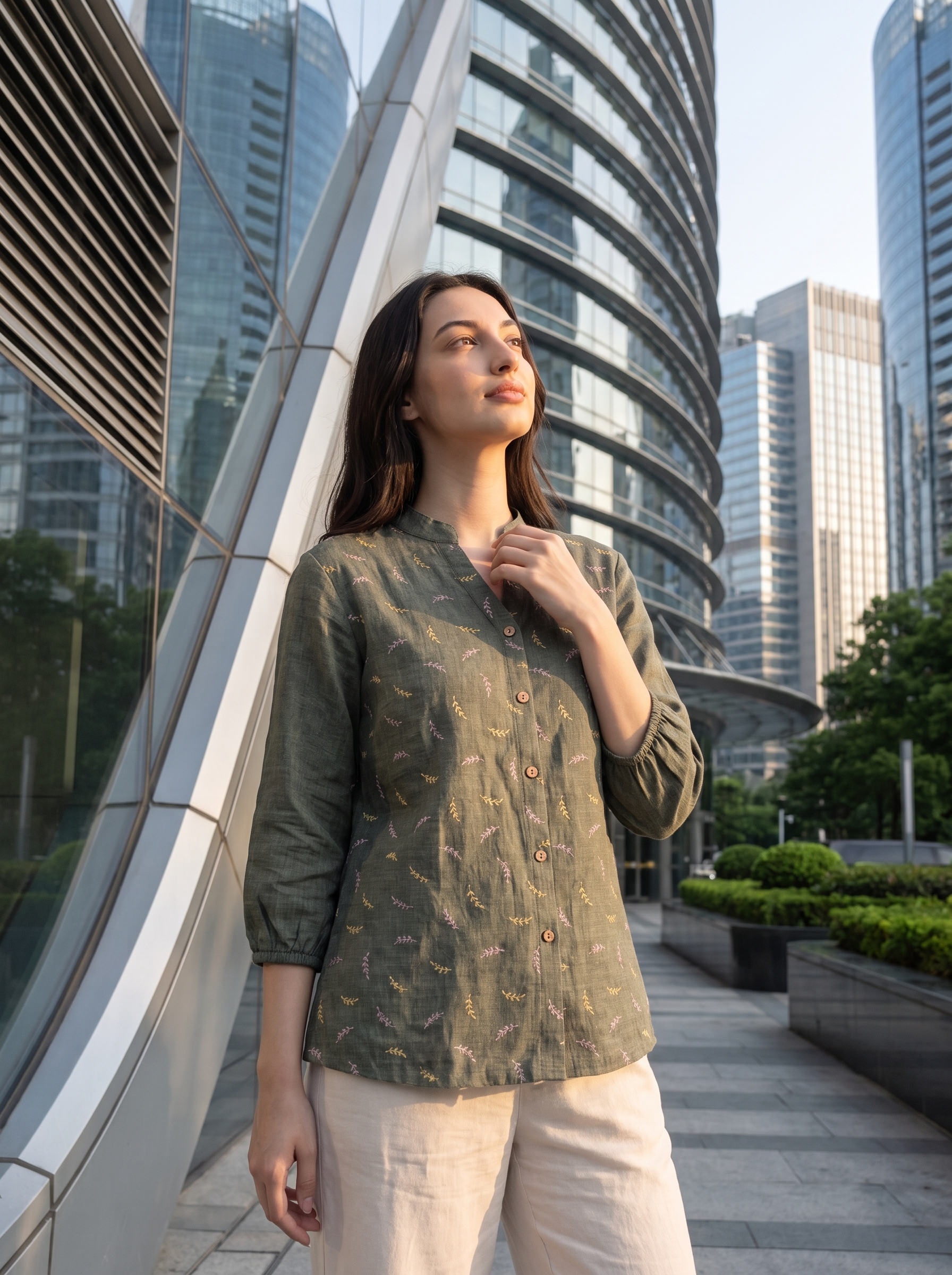 A woman wearing a sage green button-up shirt with a subtle gold leaf pattern and light beige pants, standing outdoors in front of a modern glass skyscraper.