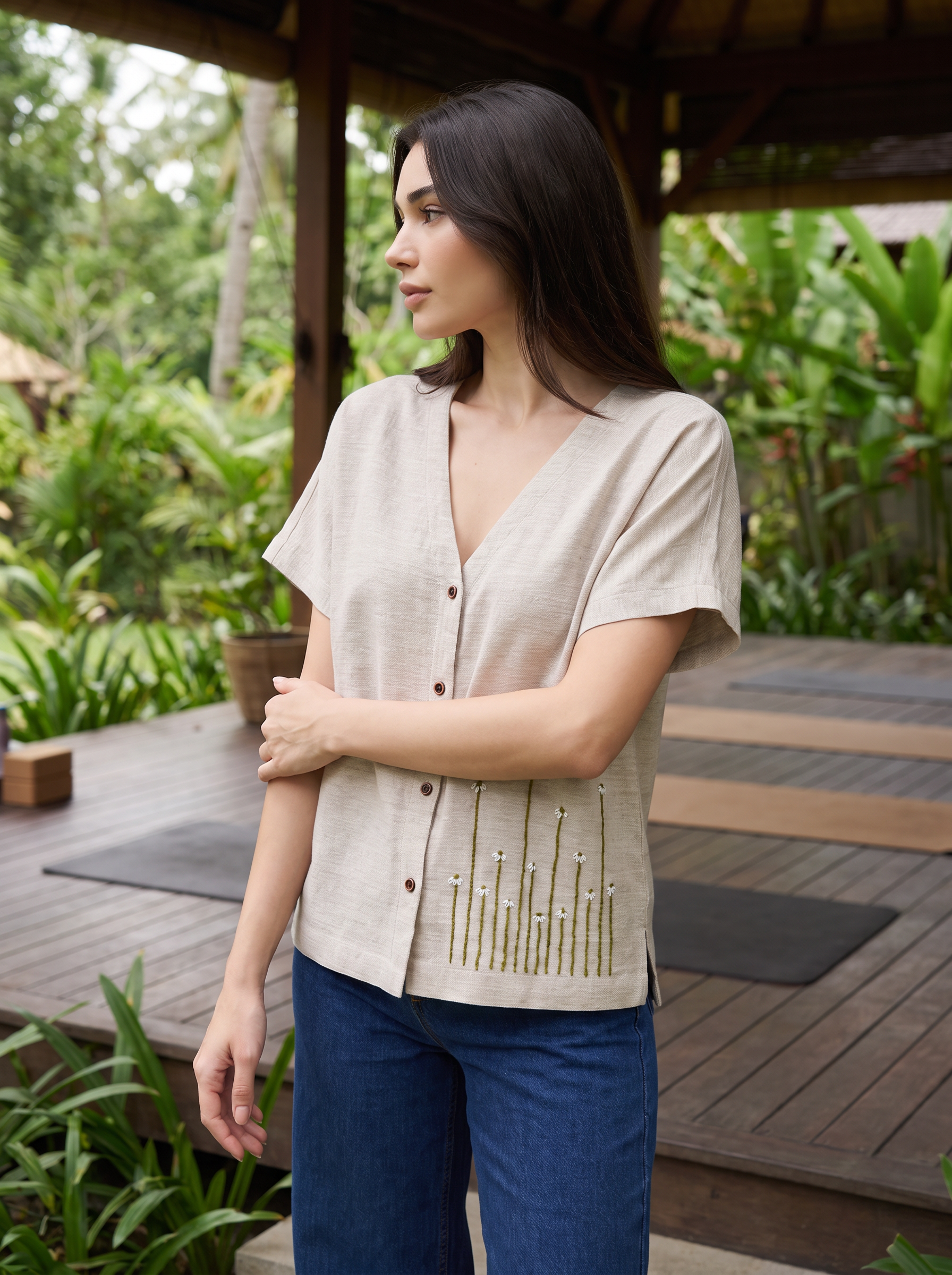 A woman wearing a light beige linen short-sleeve button-up top with gold bead embellishments on the front, paired with blue jeans, standing on a wooden deck in a lush tropical garden.