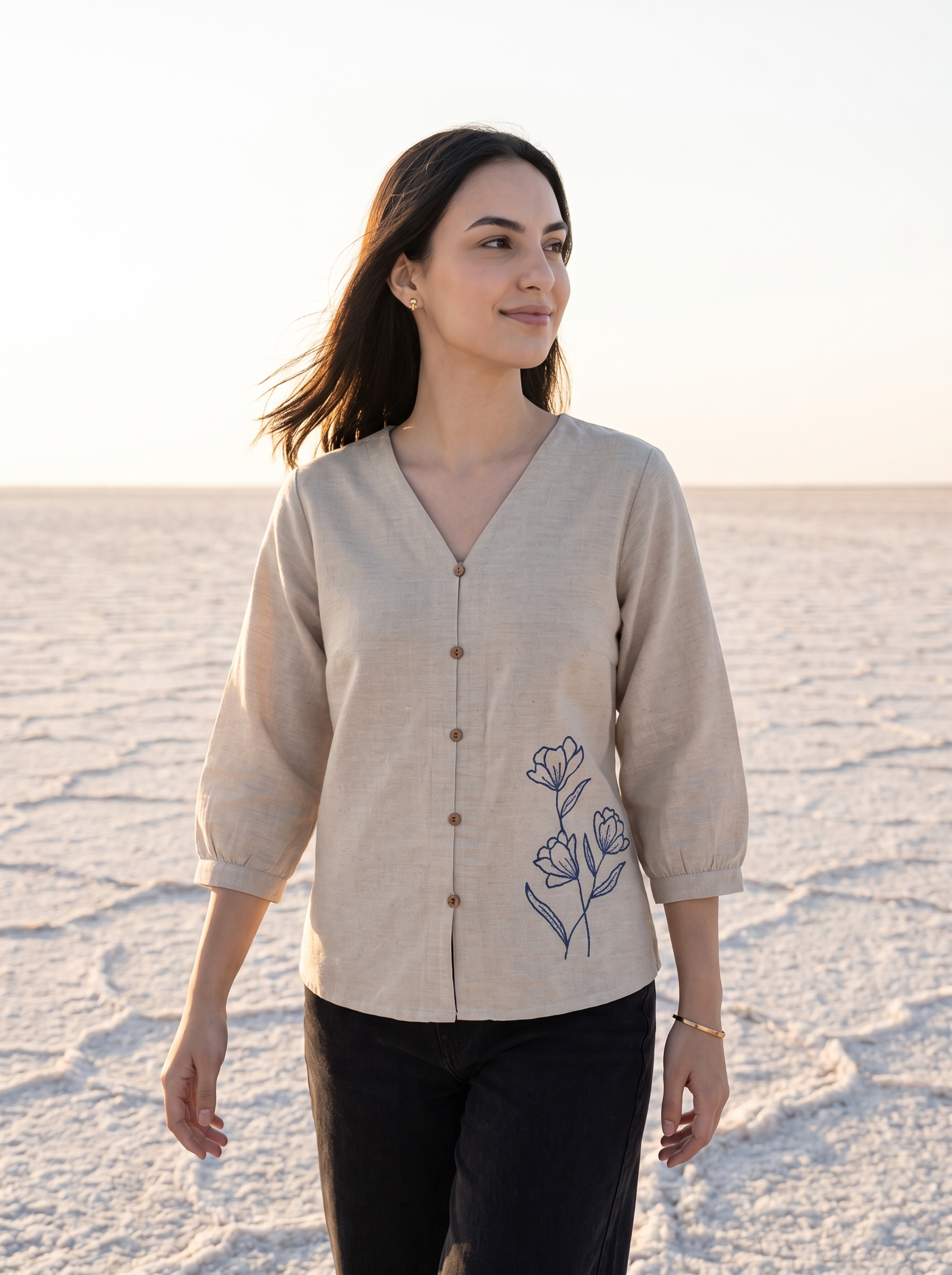 A woman wearing a beige linen button-up blouse with blue floral embroidery on the front, paired with black pants, standing on a salt flat under a bright sky.