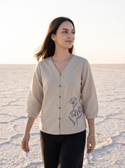 A woman wearing a beige linen button-up blouse with blue floral embroidery on the front, paired with black pants, standing on a salt flat under a bright sky.