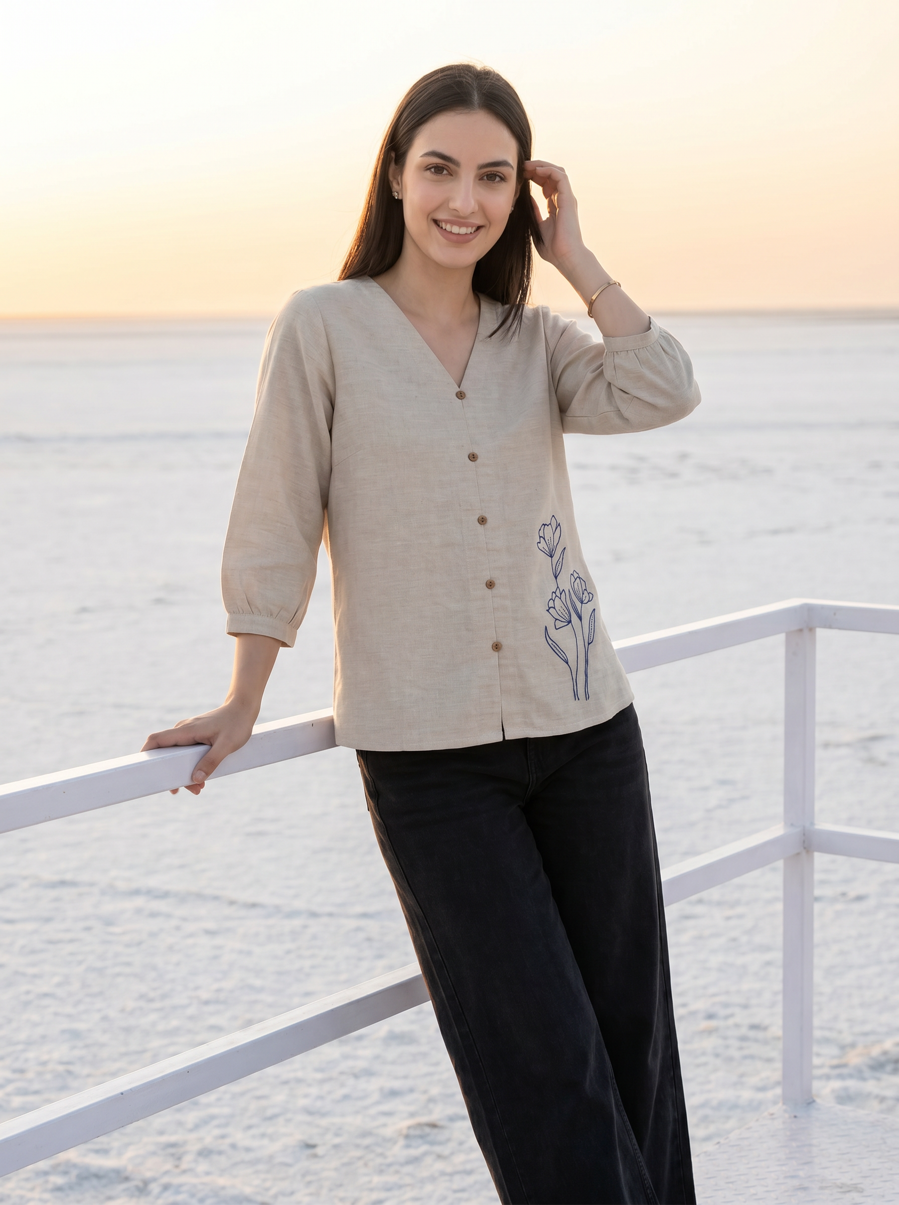 A woman stands on a white deck railing overlooking a calm ocean at sunset, wearing a light beige linen button-front top with a blue floral embroidery on the lower front and dark wide-leg pants.