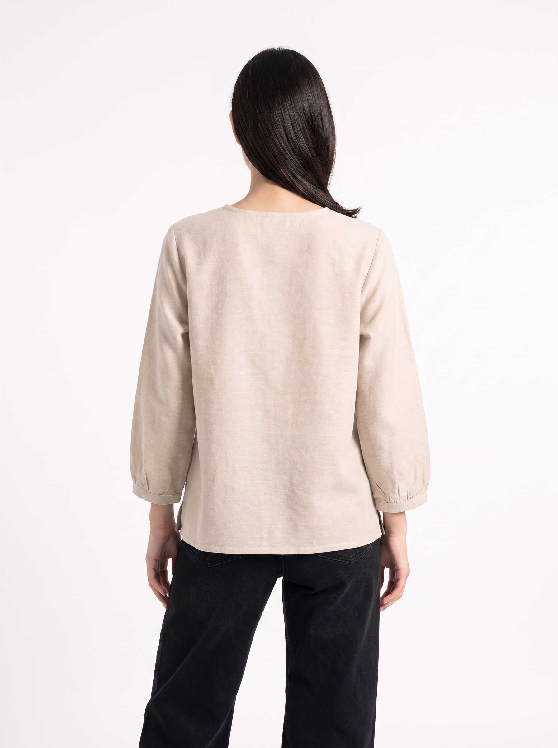 A woman viewed from the back wearing a light beige linen long-sleeve top with gathered cuffs and dark pants, set against a plain white studio background.