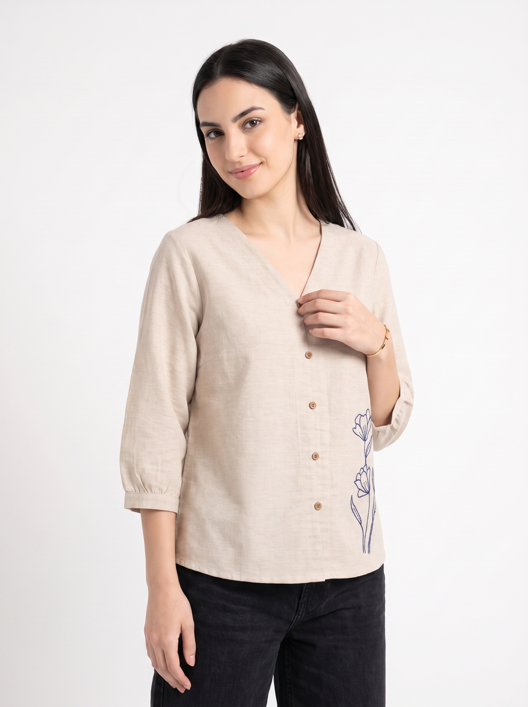 A woman modeling a light beige linen button-up blouse with a V-neck, 3/4 sleeves, and blue floral embroidery on the side, paired with black jeans against a plain white studio background.