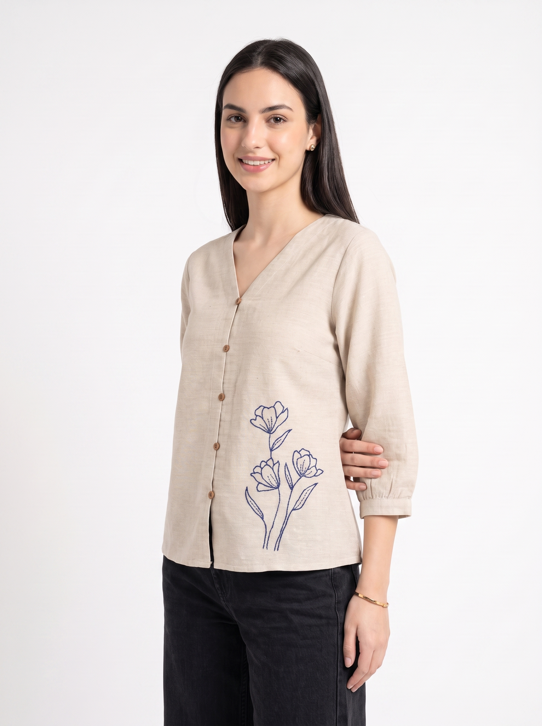 A woman modeling a light beige linen cardigan with a V-neck, brown buttons, and a blue floral print on the front, paired with black pants against a white studio background.