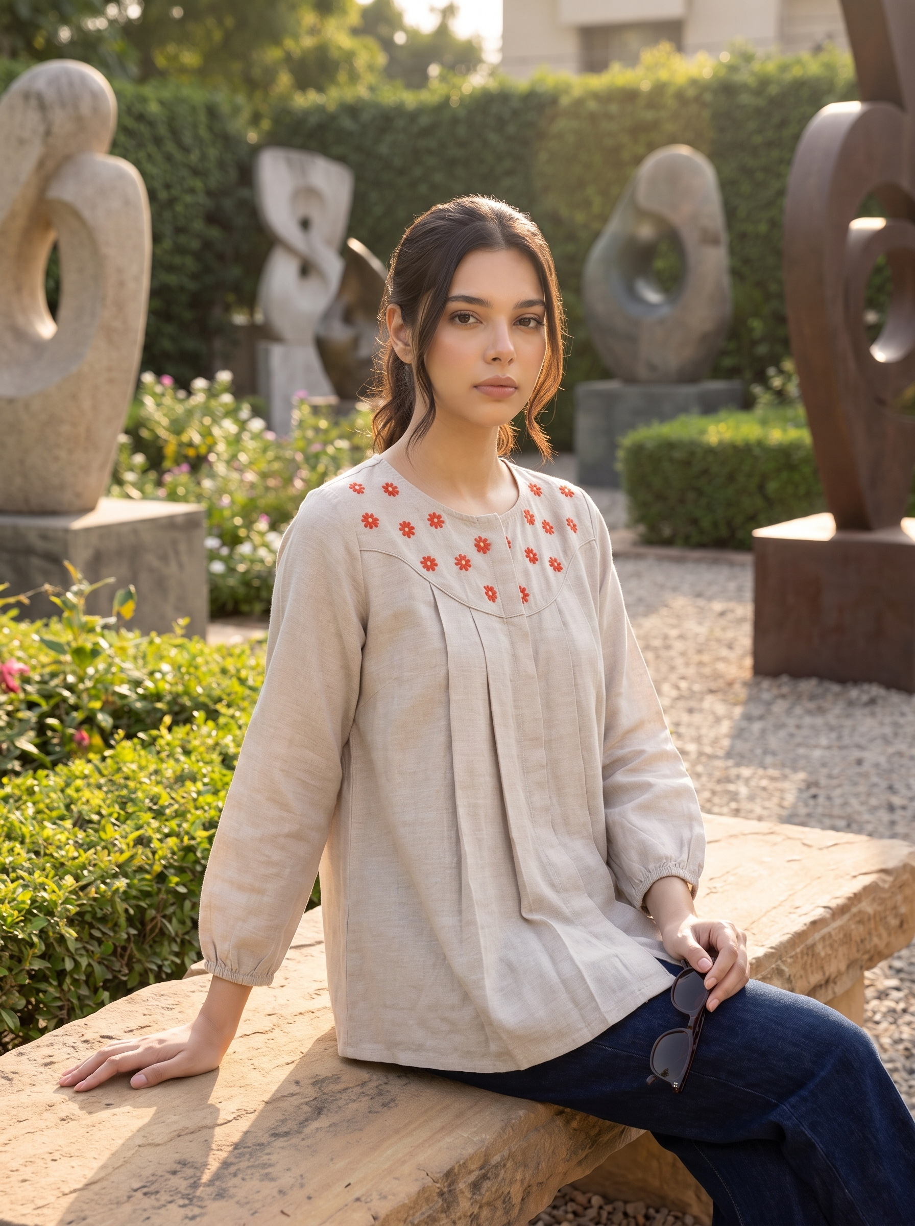 A woman sits on a wooden bench in a garden courtyard, wearing a light beige linen blouse with red floral embroidery around the neckline, paired with dark blue jeans. She holds a pair of sunglasses, with stone sculptures and green hedges in the background.