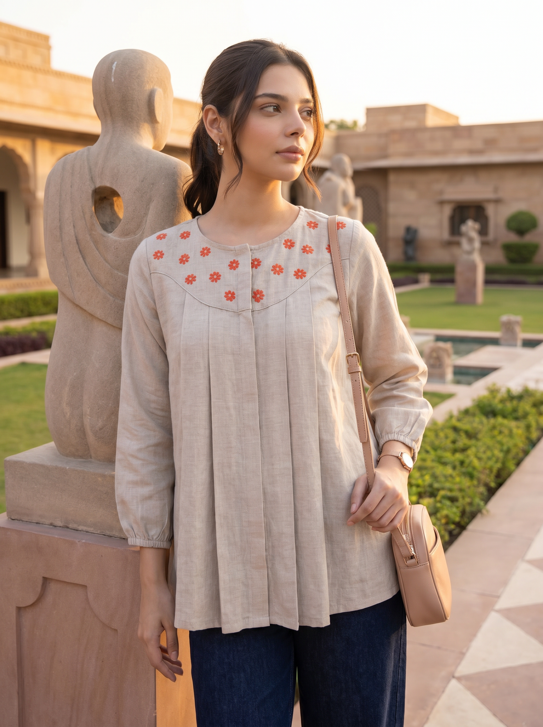 A woman wearing a beige linen tunic top with orange floral embroidery on the chest, paired with dark blue jeans and carrying a beige shoulder bag, standing in a garden courtyard with stone statues.