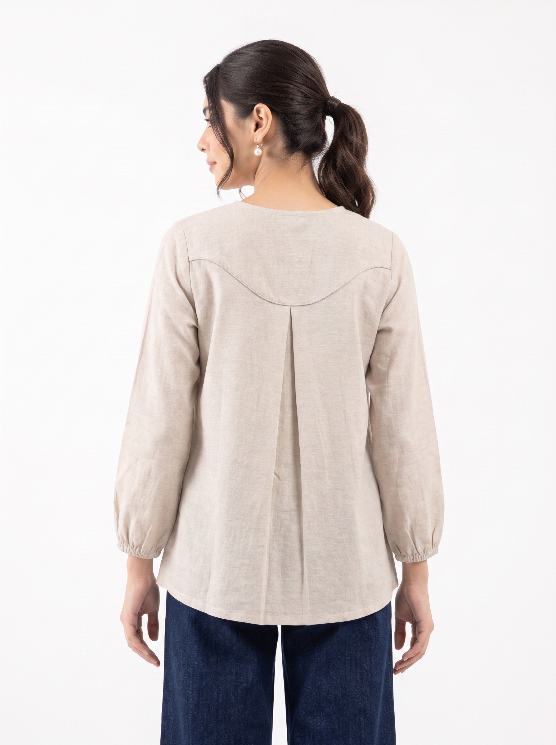 A woman shown from the back wearing a light beige linen long-sleeve top with a gathered back seam and dark blue jeans, set against a plain white studio background.