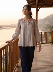 A woman stands on a wooden deck beside a body of water at sunset, wearing a light beige long-sleeve blouse with red floral embroidery around the neckline and dark navy pants.