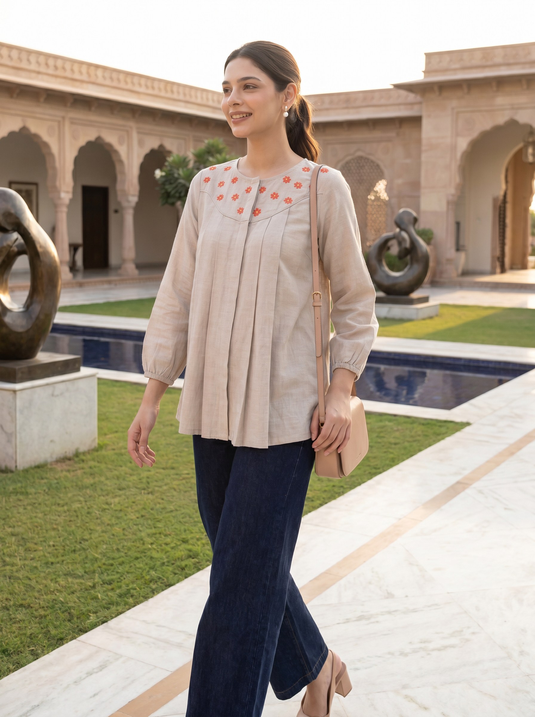 A woman models a light beige linen top with orange embroidered details on the chest, paired with dark blue wide-leg trousers, in an outdoor courtyard setting.